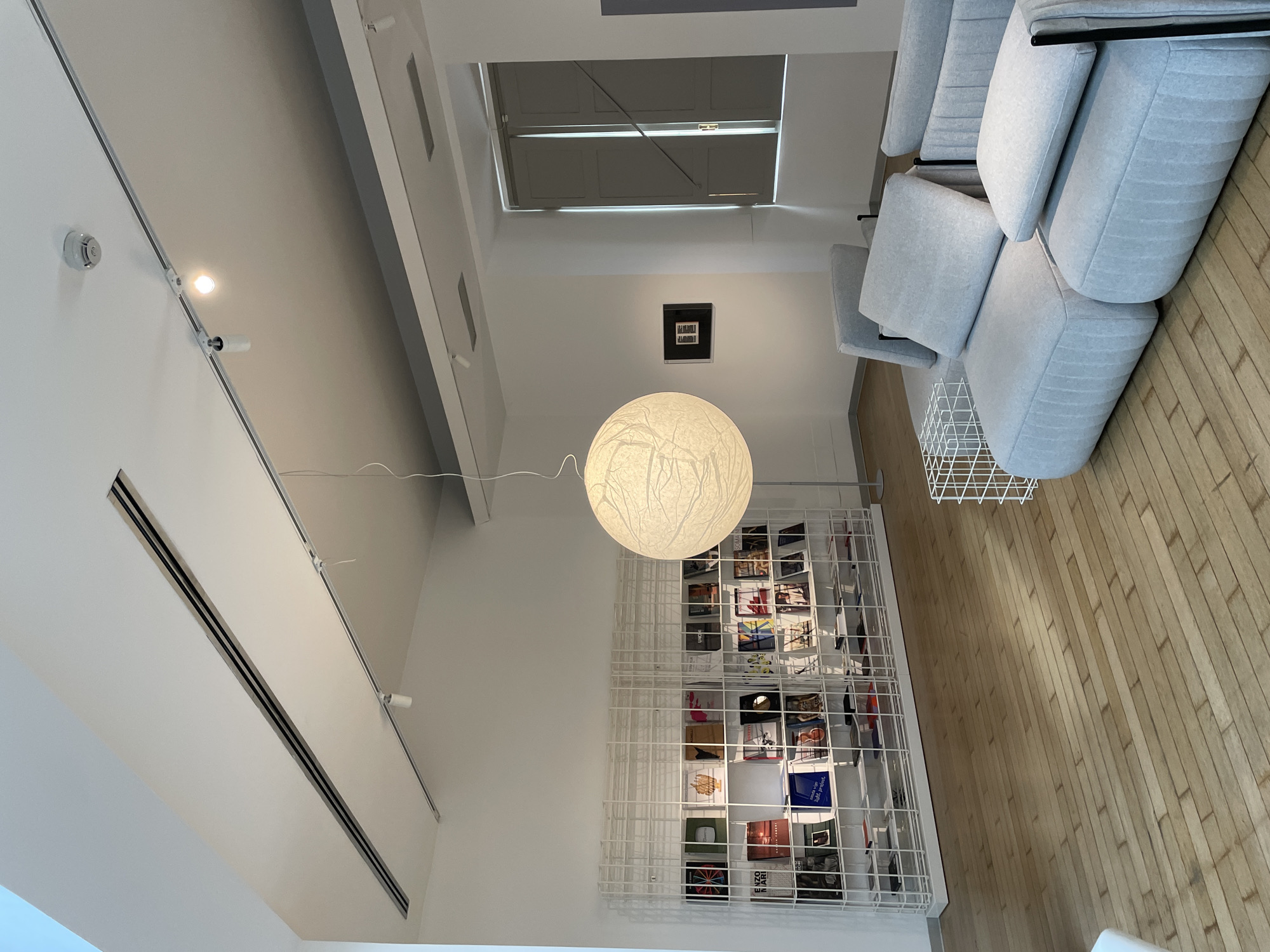 A brightly lit interior shows a room with a high ceiling in Milan, Italy. The perspective is tilted, showcasing the ceiling and the room's contents from an elevated angle. The space is characterized by modern design elements including a series of light-colored modular sofas and a tall, white wire shelving unit filled with books and magazines.
Dominating the center is a large, spherical paper lantern pendant light. The ceiling features recessed lighting tracks and vents.
The floor is made of light-colored wood planks. A small, framed picture hangs on the wall near a window, which is partially covered by a shutter or blind.
The room appears to be a showroom or lounge area, emphasizing design and aesthetic rather than function. The absence of people suggests the space is unoccupied.