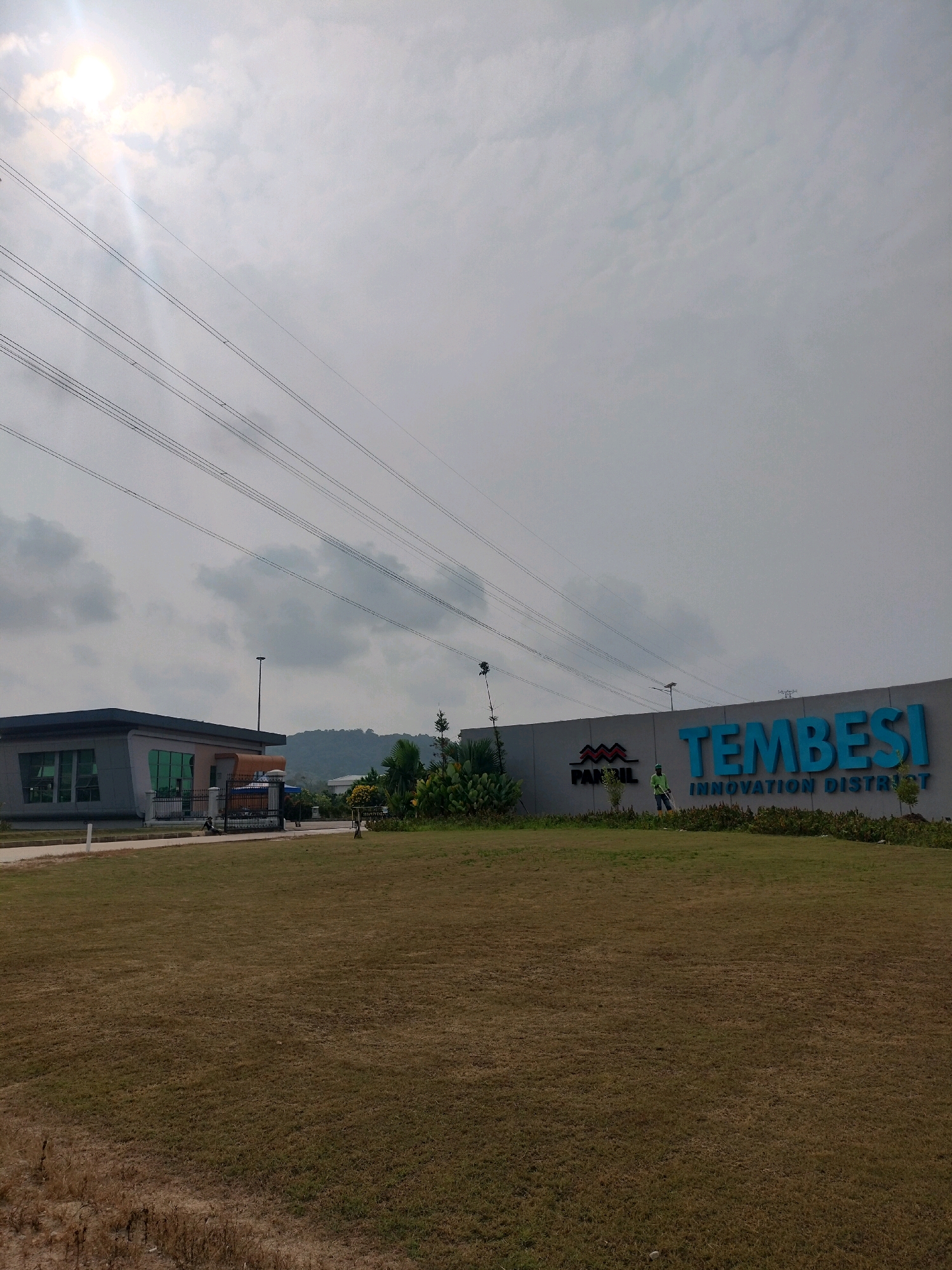 In Batam, Indonesia, the exterior of a modern, gray building with green-tinted windows is visible on the left side of the image. A black metal gate stands in front of the building. To the right, a large, curved wall displays the words "TEMBESI INNOVATION DISTRICT" in prominent blue lettering, with a smaller logo that reads "PANDIL" to its left. Two individuals, appearing to be workers, are present near the wall, one of whom is holding a tool. The foreground is dominated by a dry, brown grassy field. Overhead, multiple power lines stretch across the sky, which is partly cloudy with the sun visible. In the background, hills are discernible. The overall scene depicts an industrial or commercial area.