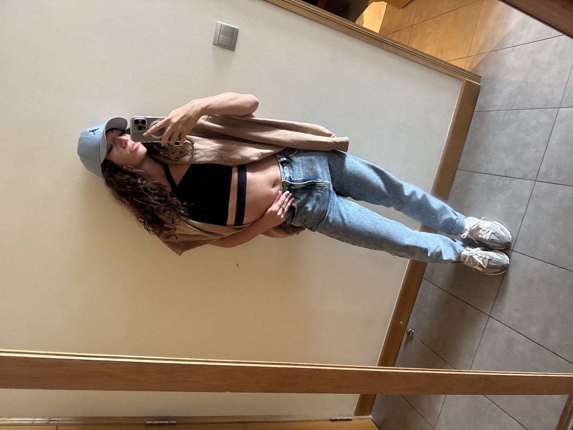 A young woman is posing for a mirror selfie in what appears to be a hotel room or apartment, based on the neutral color palette and tile flooring. She is wearing a light blue baseball cap, a black crop top with thick straps, high-waisted light blue jeans, and off-white sneakers. A tan or light brown jacket is draped over her shoulder. She is holding a smartphone in her right hand, obscuring part of her face. The room has a neutral-colored wall and tile flooring. A light switch is visible on the wall. The location of Đà Nẵng, Vietnam, cannot be confirmed from the provided image.