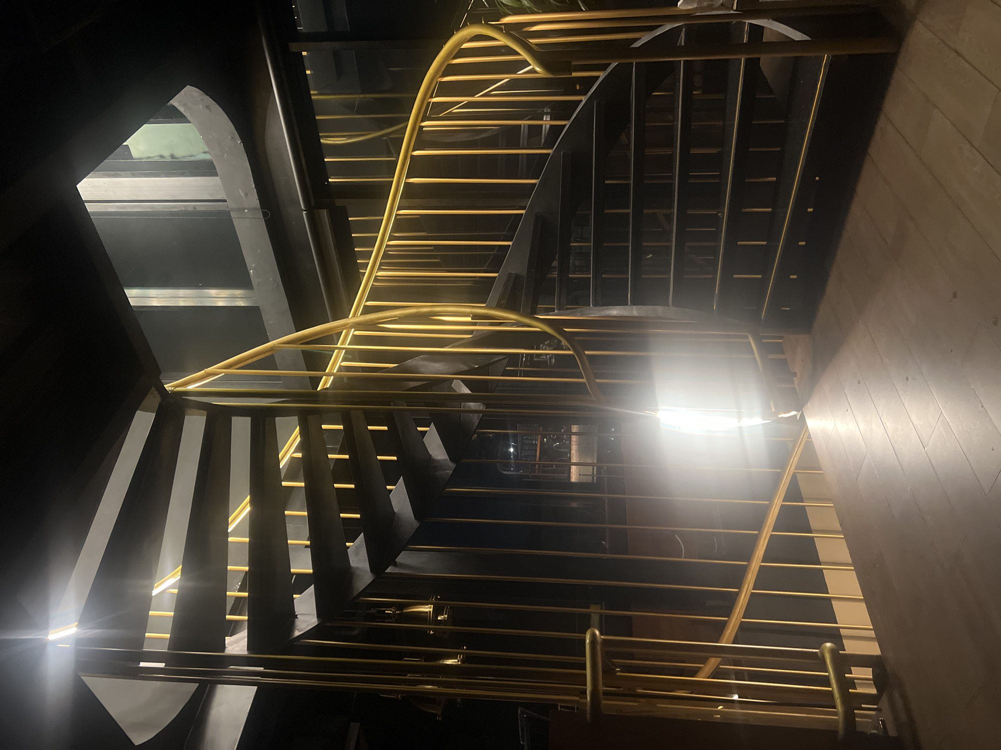 The image shows a spiral staircase with black steps and golden railings. The perspective is from below, looking up. The staircase winds around a central point. The environment is dark, but the railings are illuminated. The setting appears to be indoors, with a wooden wall visible in the background. The location is North Kuta, Indonesia.