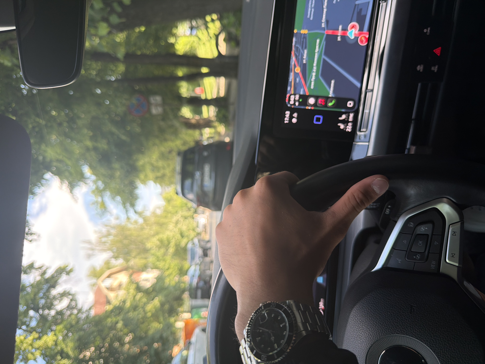 A first-person, slightly rotated, daylight shot captures a driver's perspective from inside a car in Sofia, Bulgaria. The image shows a right hand gripping the steering wheel, wearing a metallic wristwatch with a black face and bezel, resembling a luxury sports watch. The car's dashboard and a large infotainment screen are prominent, displaying a navigation map with a red route highlighting streets in Cyrillic script, specifically showing parts of "ул. Екзарх Йосиф" and "ул. Башева" which are streets in Sofia. The screen also indicates the time "17:43" (5:43 PM) and a temperature of "22.0" degrees. Below the map, several application icons are visible, along with text "GYNG" and "CLASS".
Through the windshield, a sunny outdoor scene is visible, although largely out of focus due to bokeh. Green trees line the road, and other vehicles can be vaguely seen in the distance, suggesting traffic or a street environment. A blurry road sign with a red circle and blue diagonal line (likely a "no stopping" or "no parking" sign) is also discernible. Part of the car's rearview mirror is seen in the upper left. The steering wheel features control buttons, including "OK", "VIEW", and "OFF+". The overall scene depicts a person driving or stopped in traffic during the late afternoon in an urban setting.