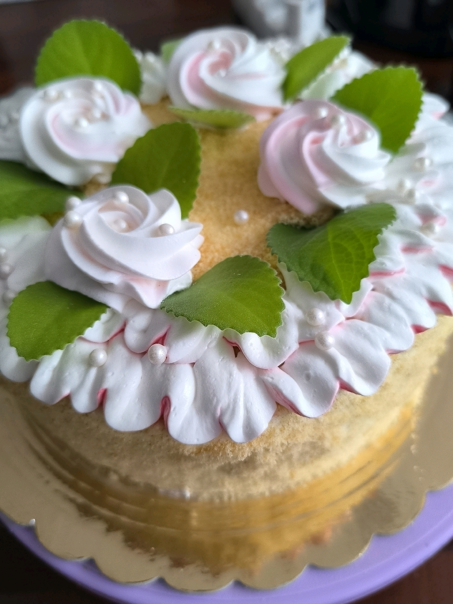 A close-up, high-angle, eye-level shot shows a round cake decorated with white and pink frosting. The cake is placed on a gold scalloped cake board, which is on a purple surface. The cake is adorned with piped frosting swirls resembling roses, some with a hint of pink. Small green leaves are interspersed among the frosting. Tiny white pearls are scattered on the frosting. The cake itself appears to be a light yellow sponge cake. The background is blurred, suggesting it is an indoor setting, possibly a kitchen or dining area. There are no people or discernible landmarks in Klaipėda, Lithuania, visible in this image.
