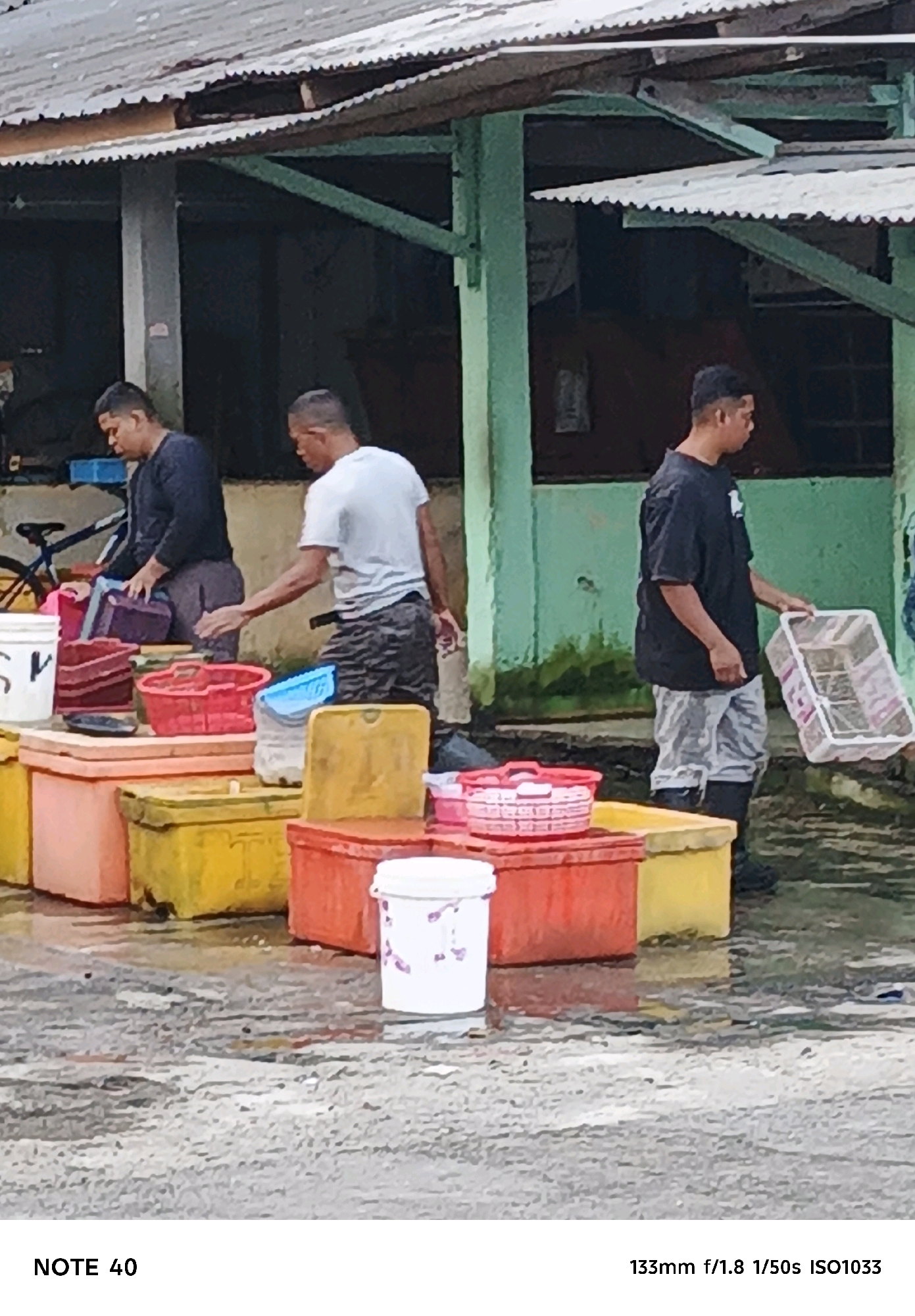 In a wet, outdoor market setting, three adult males are present. One male, wearing a dark long-sleeved shirt and grey pants, is standing and holding a white plastic crate. Another male, wearing a light-colored t-shirt and patterned grey pants, is crouched and reaching towards a collection of colorful plastic containers and coolers. A third male, wearing a dark long-sleeved shirt and dark pants, is also crouched and handling a dark purple plastic container. The ground is wet and reflective, suggesting recent rain or water spillage. A bicycle is visible in the background. The scene appears to be located in the city of Kelurahan Tegalkamulyan, Cilacap, Indonesia, as indicated by the context.