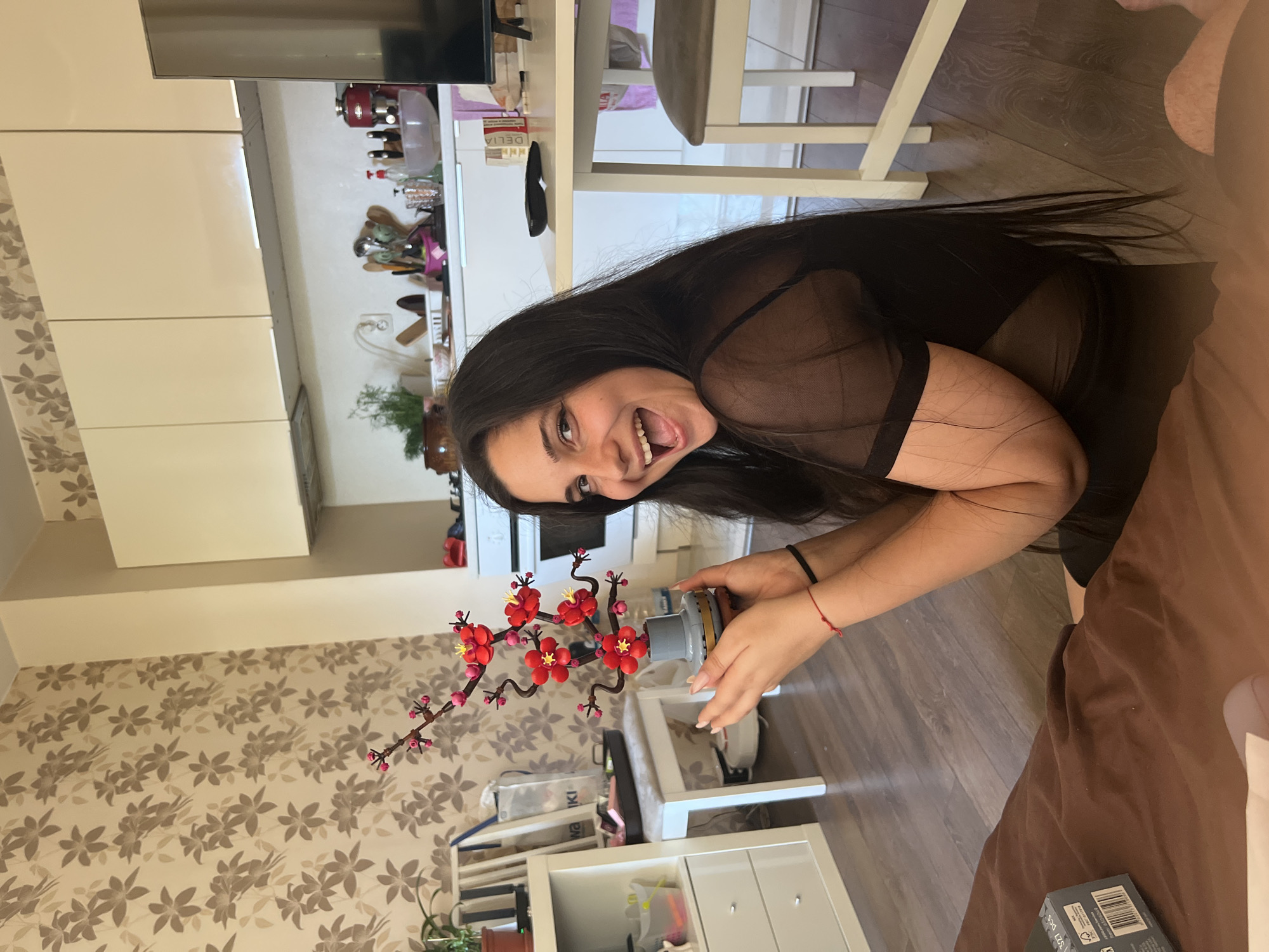 The image depicts a woman of indeterminate age, seated on the floor in what appears to be a residential kitchen. She is holding a small, decorative object, possibly a Lego build of a flowering branch, in her hands. The woman has long, dark hair and is wearing a black, sheer top. Her facial expression suggests excitement or joy, with her mouth open in a wide smile.
The environment includes a patterned wallpaper on the left wall and cream-colored cabinets and appliances in the background, characteristic of a kitchen area. A white table and chair are visible on the right side of the frame. A shelf unit with drawers and various items is present to the left of the woman. The flooring is a dark wood laminate.
The provided image does not contain any specific visual cues to indicate the location as Sofia, Bulgaria. Therefore, no details about its location in Sofia can be provided.