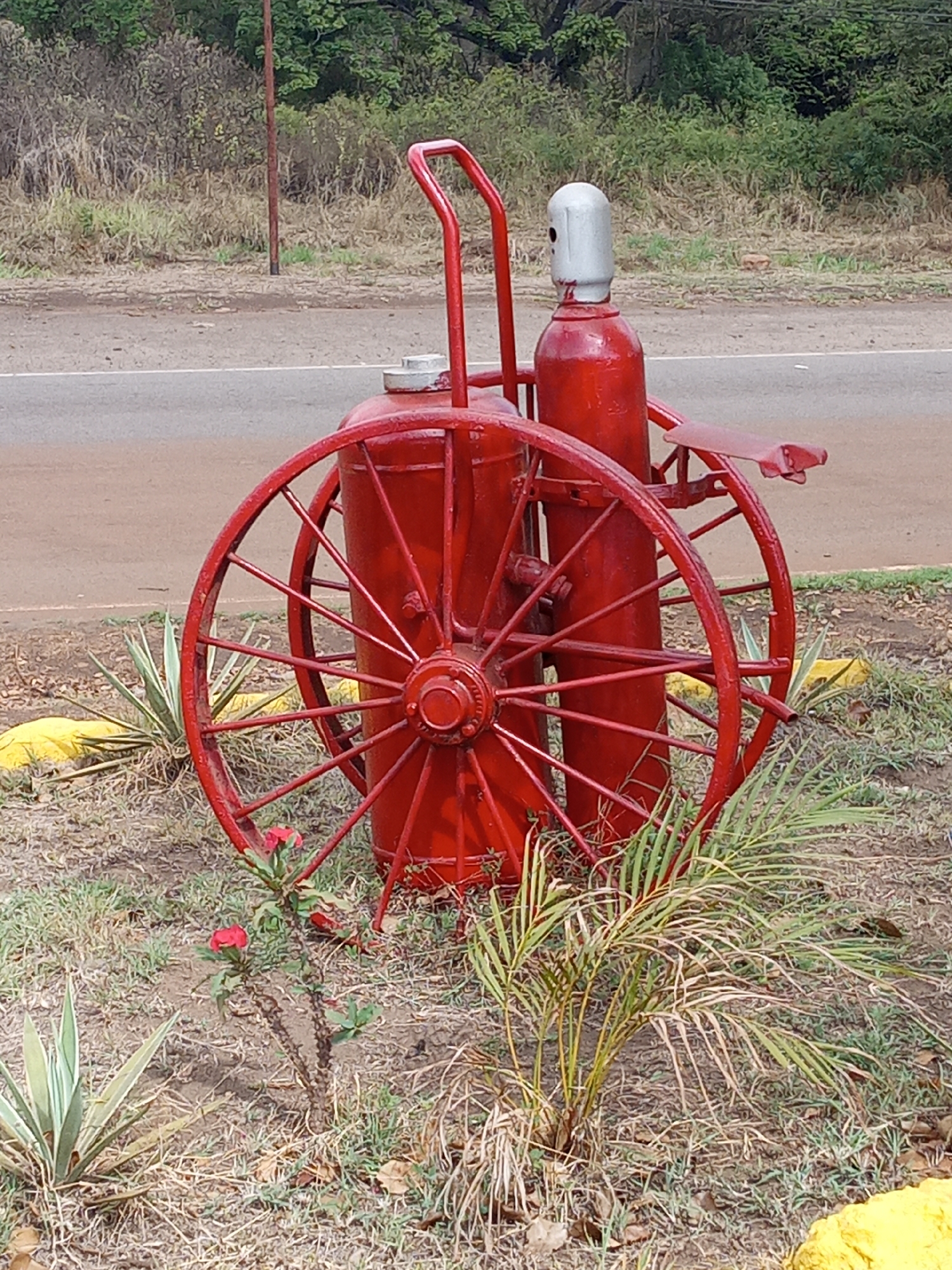 In Guayana City, Venezuela, a vintage-style red fire extinguisher apparatus stands as an ornamental piece beside a road. The device consists of two large, spoked wheels supporting a cylindrical tank and a smaller, vertical gas cylinder, all painted red except for the gas cylinder's silver top. The apparatus rests on soil covered with grass and small plants, including a small palm and a flowering shrub. A paved road with light traffic passes in the background, adjacent to a grassy embankment and shrubs.