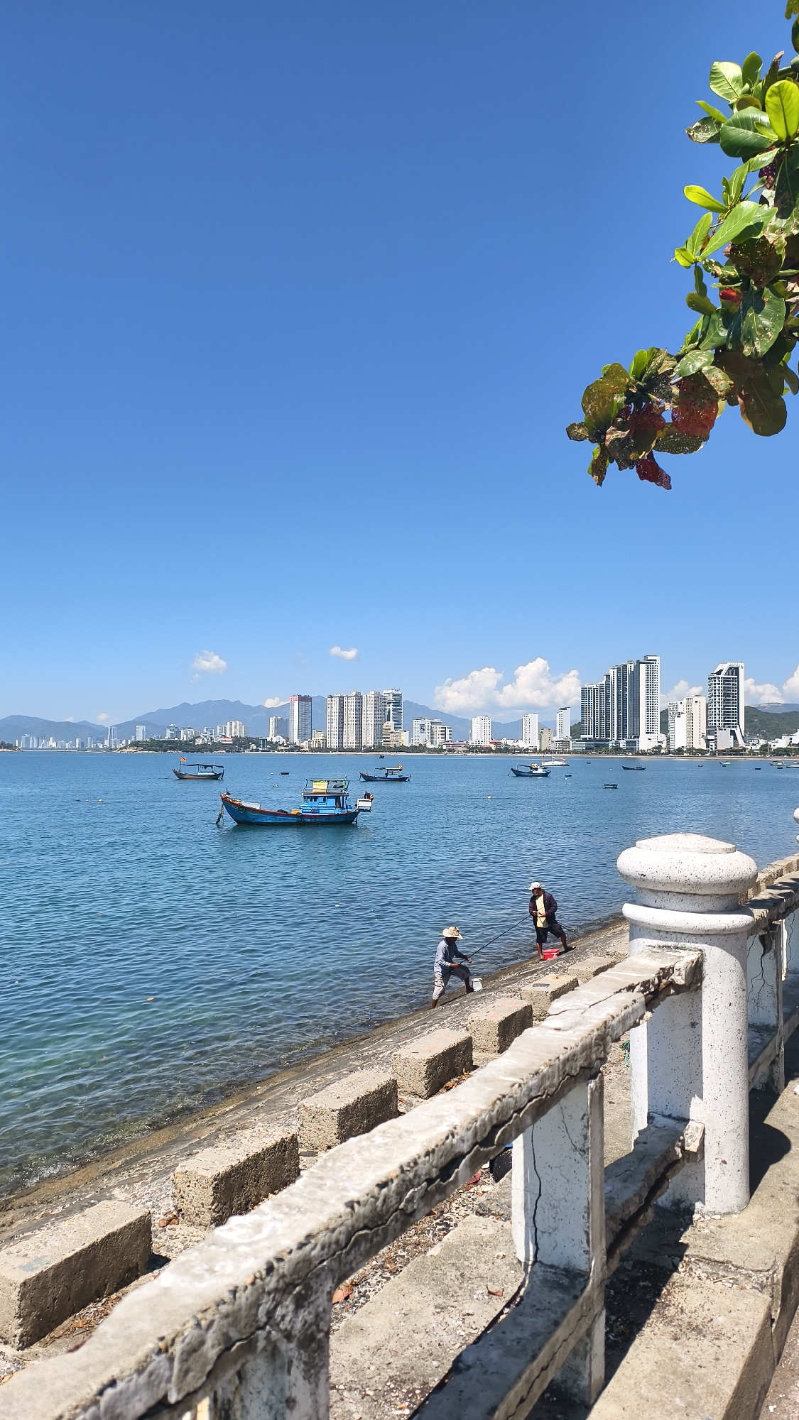 The image depicts a coastal scene in Bac Nha Trang Ward, Vietnam, under a clear blue sky. In the foreground, a weathered concrete railing lines a promenade or seawall, showing signs of age and some cracks. Beyond the railing, large concrete blocks form a textured edge to the embankment.
To the left, the calm, blue water of the sea stretches towards a distant city skyline. Several boats are visible on the water; one small blue fishing boat is closest to the shore, with others further out. The city's silhouette features numerous modern high-rise buildings and some mountains in the background.
On the shore, two individuals are engaged in fishing. One person, wearing a straw hat, is casting a fishing line into the water, while another person, also wearing a hat, stands nearby holding a fishing rod. A red bucket is placed on the embankment. Lush green foliage with some reddish leaves hangs into the frame from the top right, suggesting trees or bushes growing nearby. The overall impression is a tranquil day by the sea in a developing urban coastal area.