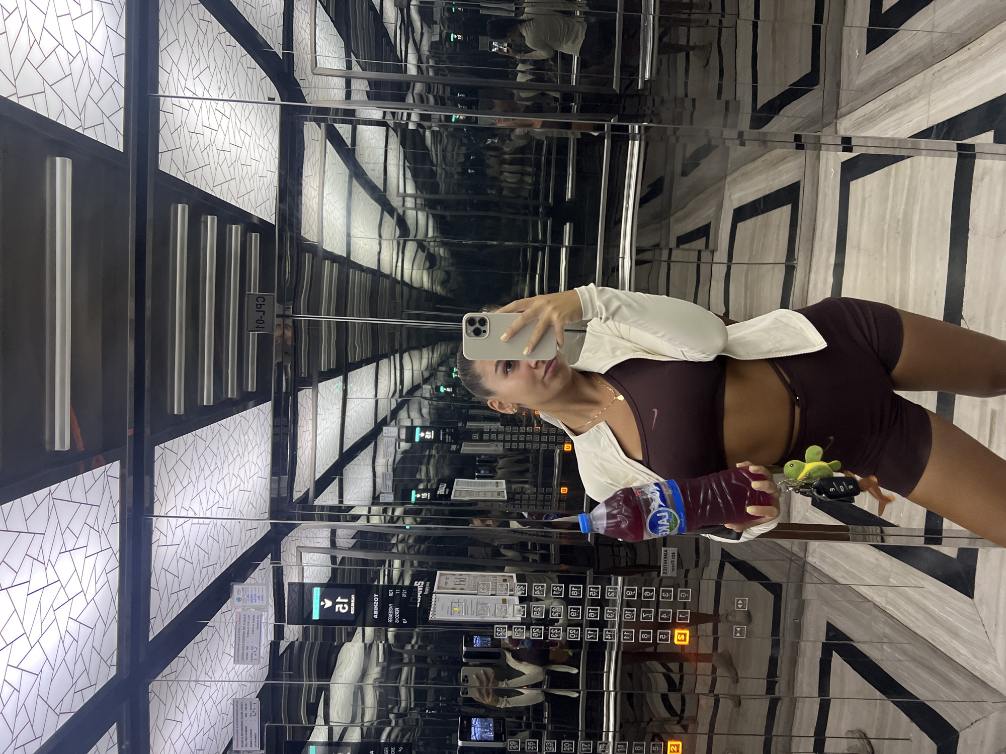 A young woman takes a selfie in an elevator, with the doors covered in mirrors, creating multiple reflections and repeating patterns. She is wearing a dark athletic bra and matching shorts with a white jacket tied around her waist. She holds a bottle of juice in one hand and her car keys with a turtle keychain in the other. The elevator's buttons are visible, with several lit up, and signs are posted in both English and another language. The elevator's interior features geometric designs and silver accents. The OCR indicates some of the labels on the elevator, including the floor numbers and elevator functions.