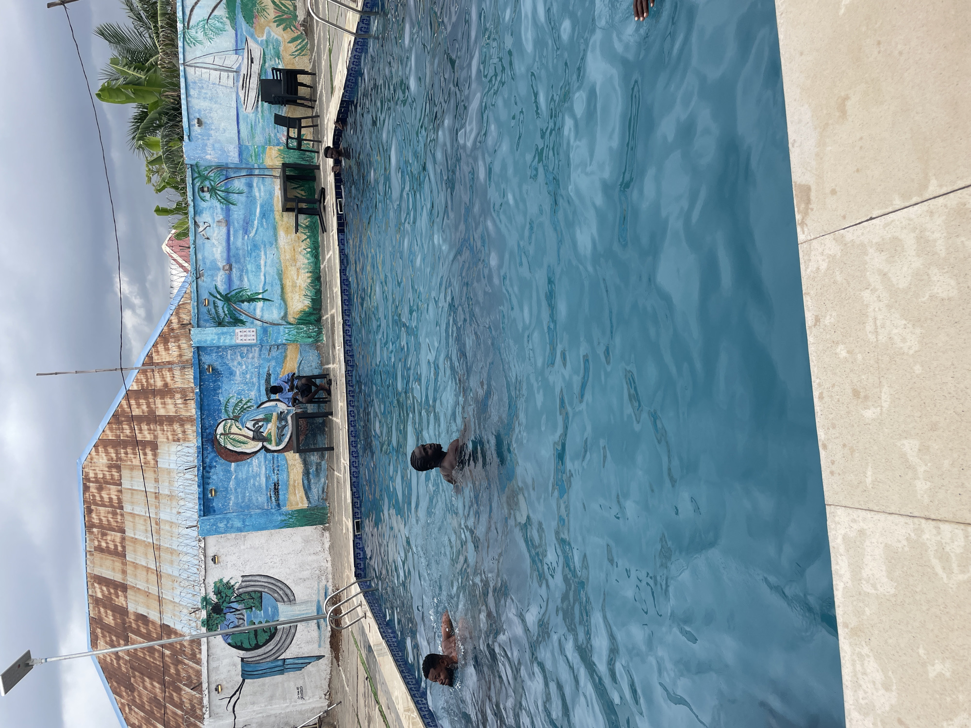 The image depicts a swimming pool at an outdoor location. Two people are swimming in the pool. The pool is situated next to a wall with mural paintings of beach scenes and palm trees. There are chairs and tables alongside the pool. The background shows a building with a corrugated metal roof and barbed wire along the top of the wall. The sky appears overcast. While I cannot confirm the location, Akwanga, Nigeria is given as the city.
