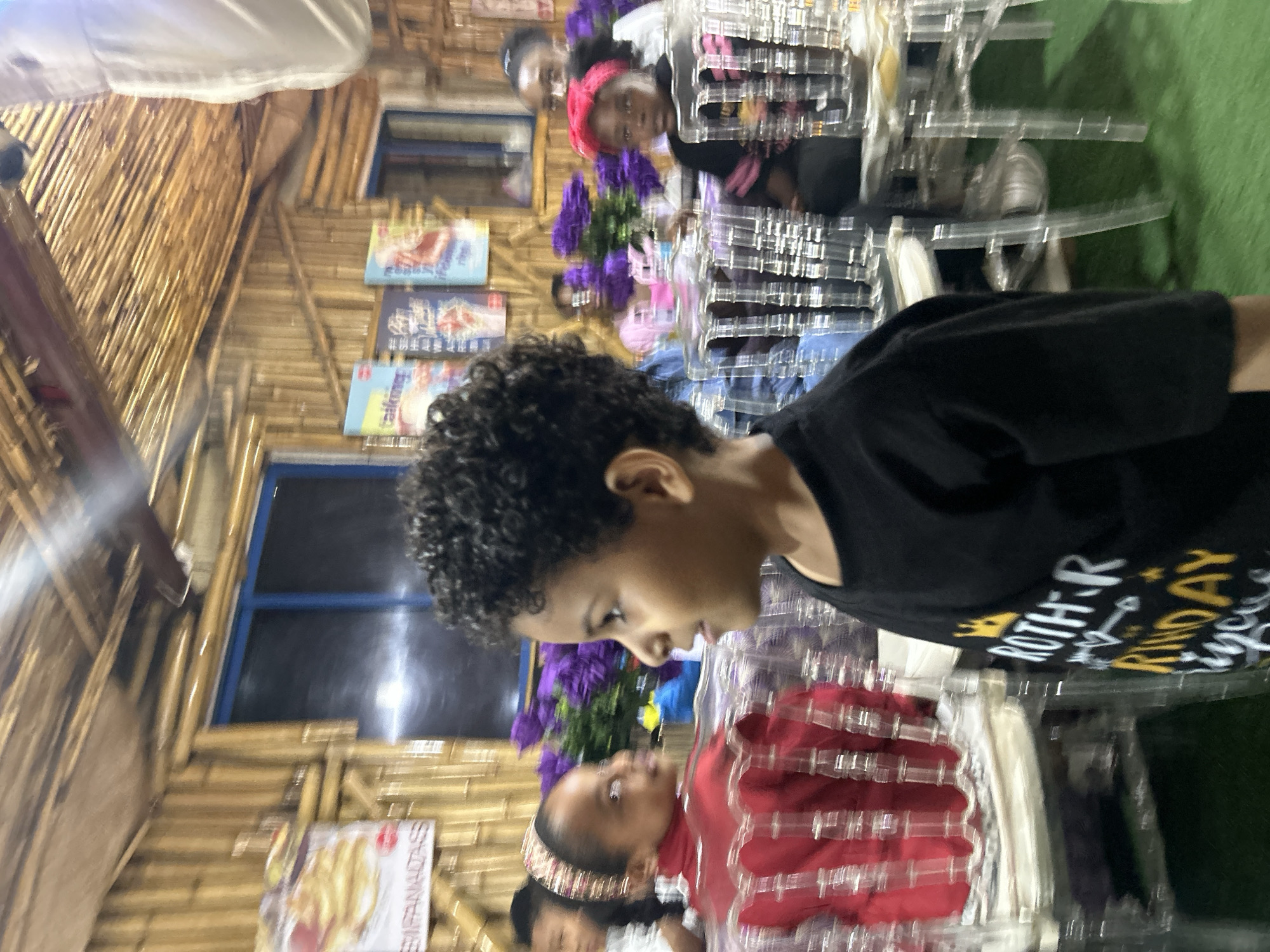In Bar Jirgi Summa, Nigeria, this image captures a dimly lit interior, likely a restaurant or event space, decorated with bamboo walls and ceiling. The lighting is soft and warm, emanating from above and from behind the subjects, creating a somewhat hazy atmosphere. In the foreground, a young boy with curly hair is the primary subject, facing to the right, with his mouth slightly open as if speaking. He is wearing a black t-shirt with yellow and white lettering that appears to read "ROTHR KINDAY," suggesting a birthday celebration. To his left and slightly behind him, a young girl in a red top and a decorative headband is also looking to the side. Other children and adults are visible in the background, some seated at tables with clear plastic chairs, and purple floral arrangements add a touch of color to the scene. The overall impression is of a casual gathering, possibly a birthday party, taking place in a unique, rustic-themed venue.