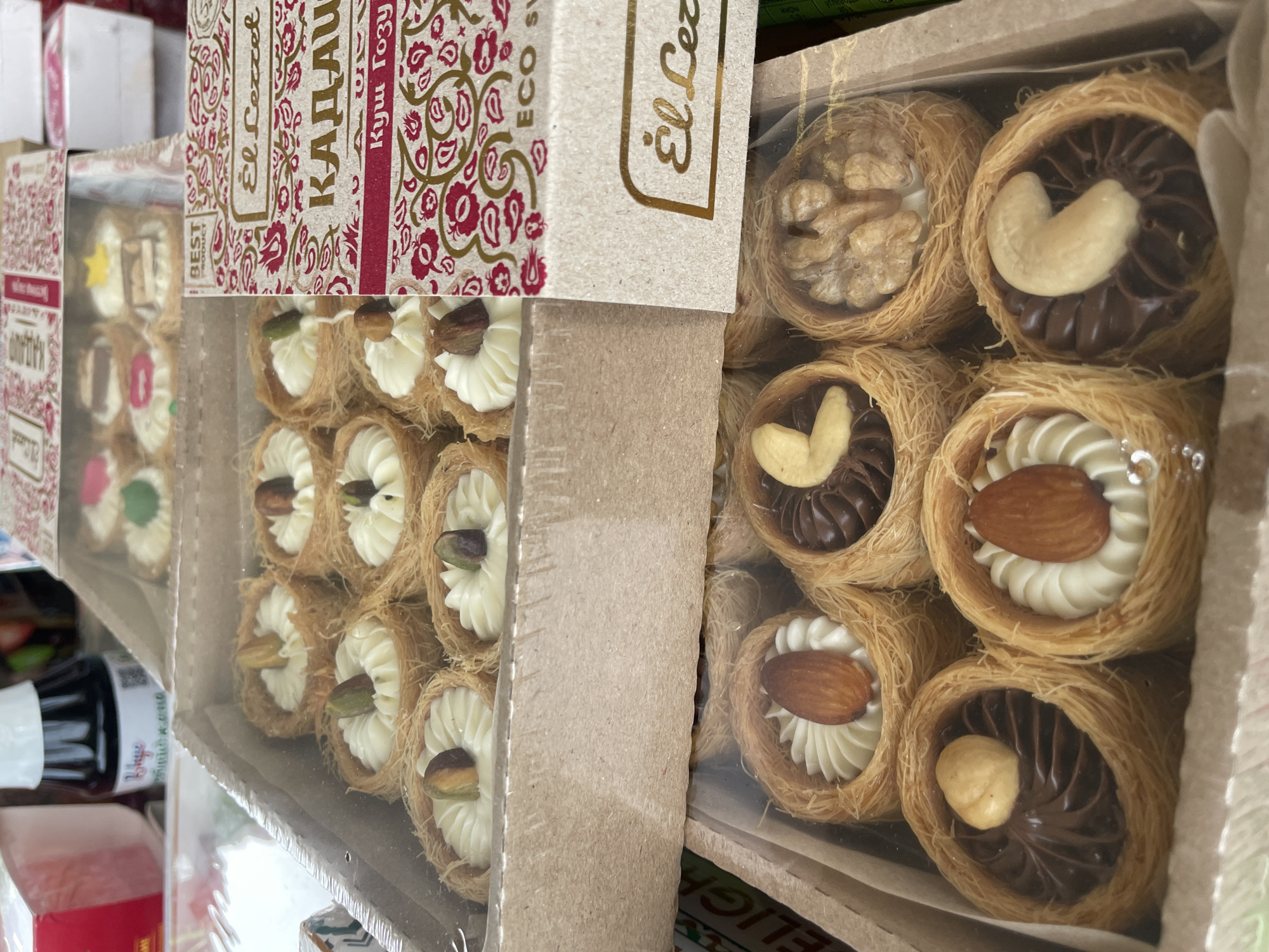 The image shows two boxes of what appear to be Turkish delight or similar types of confectionary, likely photographed indoors at a store or market. The box on the left contains pastries filled with white cream and topped with a pistachio. The box on the right contains pastries filled with chocolate and topped with either a cashew or almond. The packaging bears the brand name “El Lezzet” and the phrase “Eco Sweet”. “КАДАИФ” is visible in large letters on the boxes. The location is Olginka, Russia.
