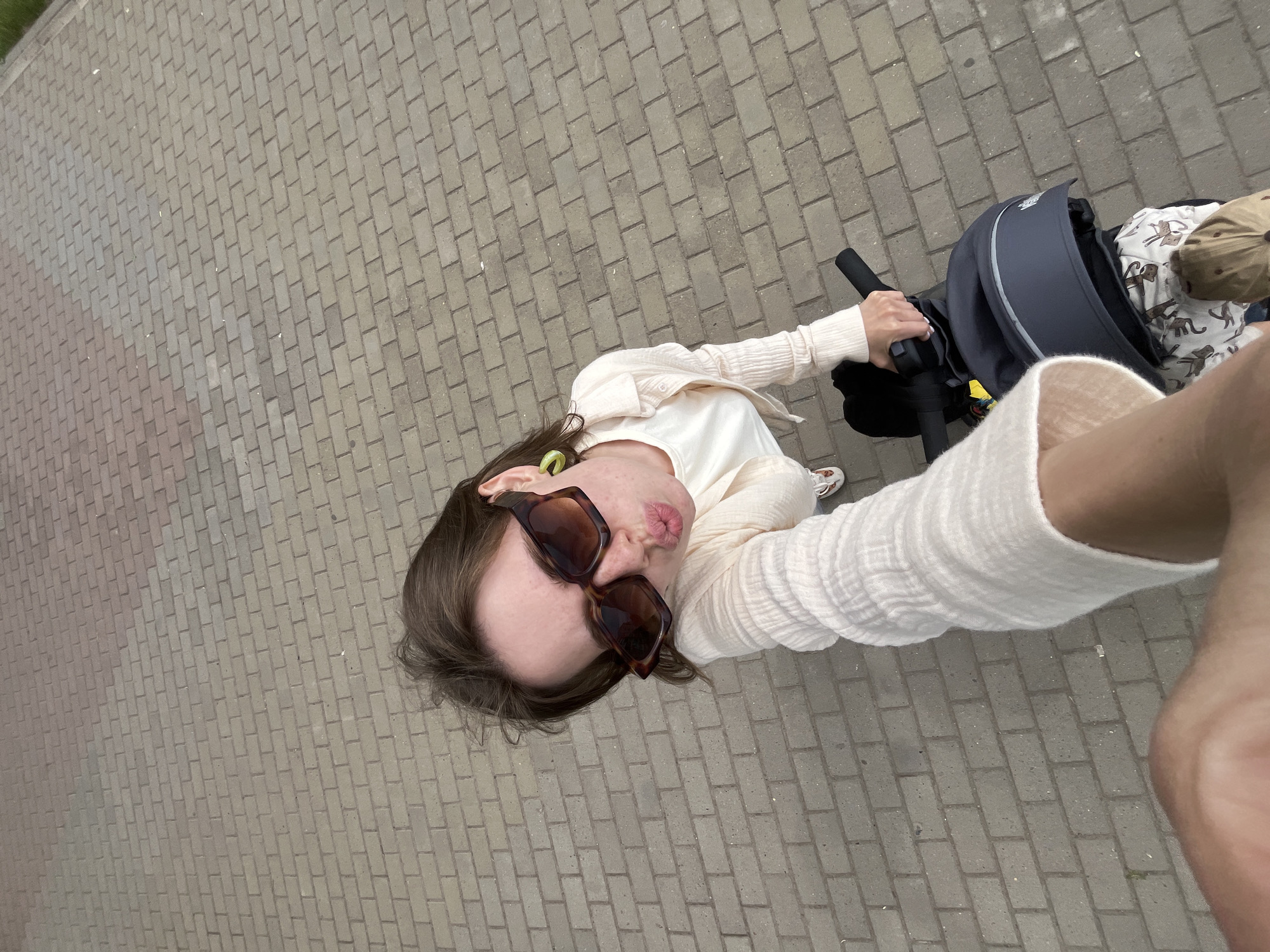 In an overhead, wide-angle shot, a young Caucasian woman is standing on a brick sidewalk in Chaikovsky, Russia. She is wearing sunglasses, a light-colored cardigan over a white top, and is making a kissing gesture. She is holding onto the handlebars of what appears to be a stroller or a similar wheeled device. The ground is covered in light-colored bricks, and there is a black bag on the stroller. The woman has brown hair and light skin, and she is wearing green earrings. The background is a brick walkway extending into the distance.