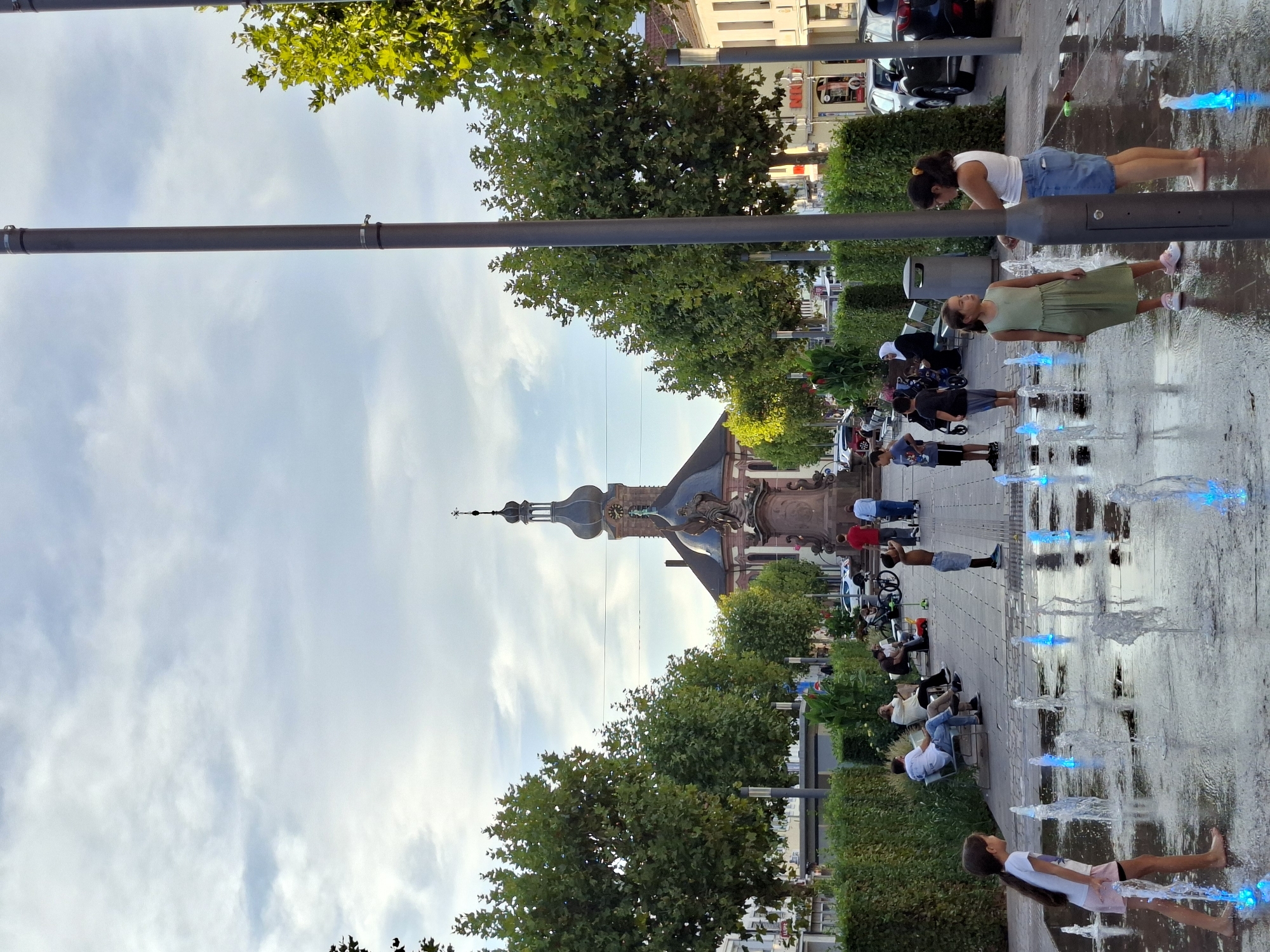 The image depicts a public square in Rastatt, Germany, with a prominent church and a statue at its center. In the foreground, children are playing in a water fountain with illuminated blue jets. Several adults are seated on benches in the background, observing the scene. The square is lined with trees and hedges, and there are buildings and parked cars on either side. The sky is cloudy, indicating overcast weather. The overall impression is that of a peaceful, community-oriented space where people are enjoying a day outdoors.