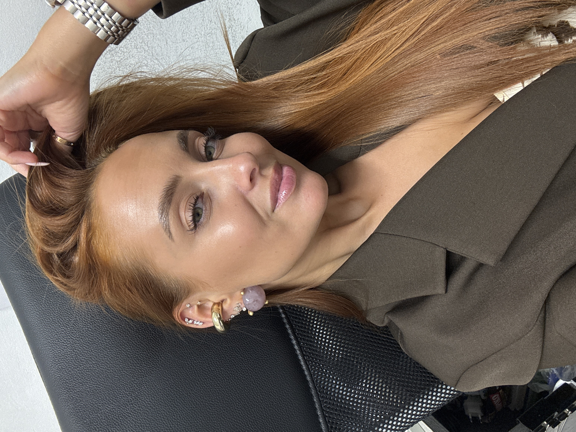 The image presents a close-up portrait of a young adult woman with long, reddish-brown hair and light green eyes, lying down and looking towards the camera with a slight, calm smile. She wears visible makeup, including lip gloss, eyeliner, and mascara. Her head rests on a dark, possibly black, leather or synthetic material surface, which appears to be part of a chair, with a black mesh section also visible.
She is wearing a dark olive-green or dark brown blazer or jacket. Her jewelry includes a silver-toned, rectangular-linked watch on her left wrist and a thin gold-toned ring with an indistinct inscription on her left ring finger. Her right ear is adorned with multiple earrings: a large, spherical light purple stone earring on the lobe, a gold-toned ear cuff, and several small, sparkling stud earrings arranged along her ear's cartilage. Her left hand, with manicured nails, is raised and lightly touching her hair.
The background shows a light-colored, textured wall in the upper left, suggesting an indoor setting. The image conveys a relaxed and posed moment. The provided location context for this media file is Chaikovsky, Russia.