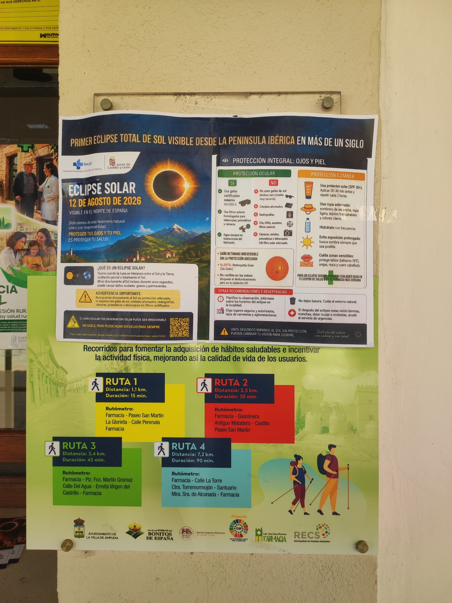 The image is a poster promoting the observation of a total solar eclipse visible from the Iberian Peninsula on August 12, 2026, specifically in the north of Spain. The poster is located in the city of Ampudia, Spain, as indicated by the logos of "AYUNTAMIENTO DE LA VILLA DE AMPUDIA" and "BONITOS DE ESPAÑA" at the bottom left.
The poster is divided into several sections. The top section features a large graphic of a solar eclipse, with the text "PRIMER ECLIPSE TOTAL DE SOL VISIBLE DESDE LA PENÍNSULA IBÉRICA EN MÁS DE UN SIGLO" above it. Below the eclipse graphic, it states "ECLIPSE SOLAR 12 DE AGOSTO DE 2026 VISIBLE EN EL NORTE DE ESPAÑA" and a tagline encouraging responsible observation.
A significant portion of the poster is dedicated to safety guidelines for observing the eclipse, with sections on "PROTECCIÓN OCULAR" (ocular protection) and "PROTECCIÓN CUTÁNEA" (skin protection). It lists recommended and not recommended practices for eye and skin safety. There is also a section titled "ADVERTENCIA IMPORTANTE" (important warning) emphasizing the irreversible damage of looking at the sun without proper protection.
The lower half of the poster presents four walking routes within Ampudia, labeled "RUTA 1" through "RUTA 4," with their respective distances and durations. Each route has a "Rutómetro" listing points of interest, primarily pharmacies and other landmarks. Illustrations of hikers are placed near these route descriptions.
The poster is attached to a beige-colored wall, with a metal stud visible in the bottom center holding it in place. The overall scene depicts a public information poster displayed outdoors.