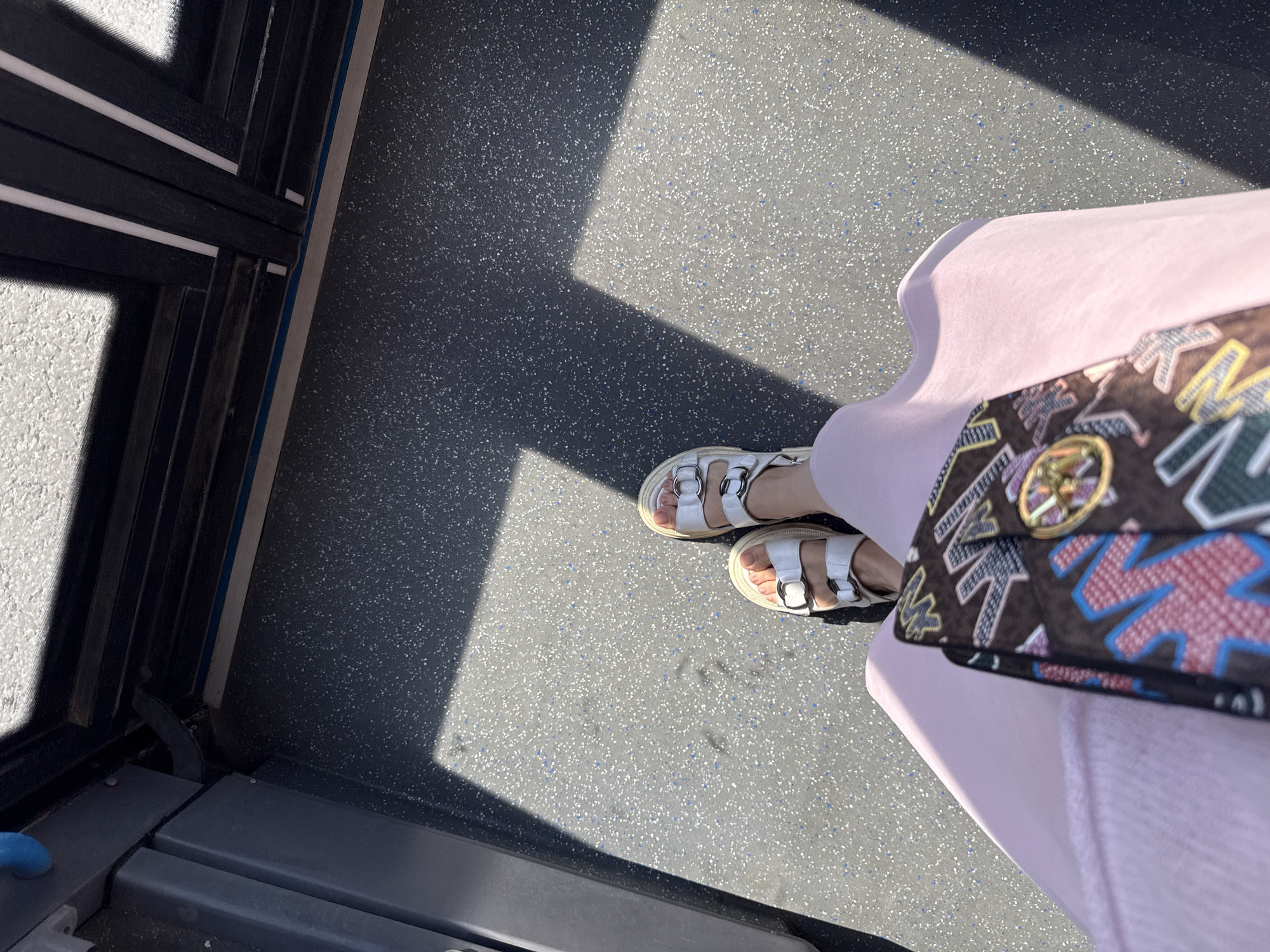The image captures an overhead view of a person's lower body and feet within what appears to be a public transportation vehicle in Moscow, Russia. The person is wearing light pink clothing, likely a skirt or dress, and white, chunky sandals with multiple straps and silver-toned buckles on their feet. A patterned bag, adorned with colorful, stylized block letters (such as green, blue, and red) and a circular golden emblem, is partially visible, resting on the pink garment.
The vehicle's floor is a dark gray with scattered light gray and blue speckles, typical of a durable, non-slip surface found in buses or trams. Bright sunlight streams in from the left, casting distinct, angular shadows across the floor, indicating a sunny daytime. To the left, dark metal frames and panels, consistent with a door or window structure of public transport, are visible. A thin blue line runs along the floor's edge beside these metal components. Beyond the vehicle's interior, a lighter, textured surface, possibly an outdoor environment, is faintly seen.
The scene depicts the person in transit, presumably standing or sitting. No specific emotions are discernible. The stylized letters on the bag are not clearly legible.