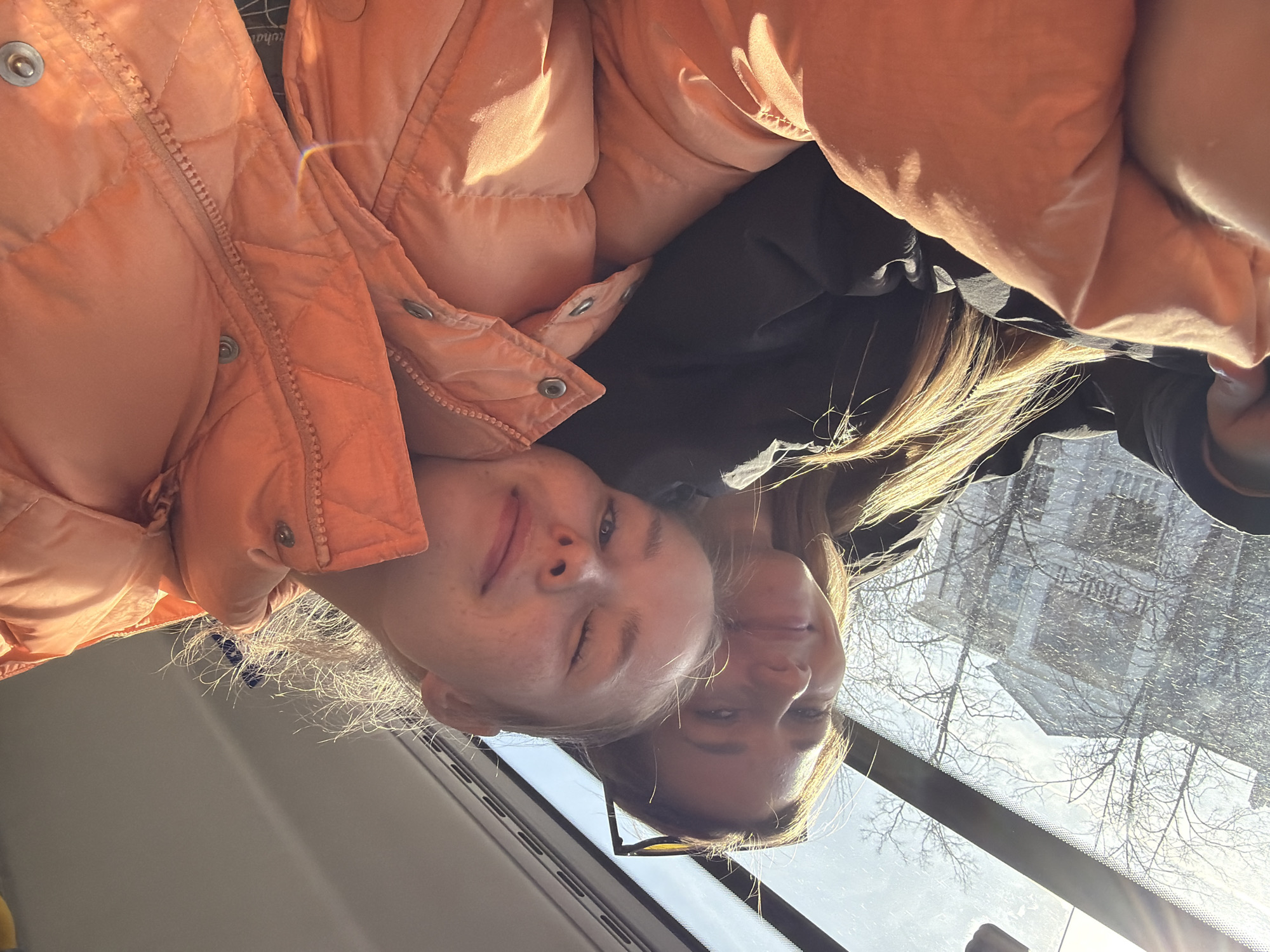 Two young women are visible from the chest up, lying side-by-side and facing the camera. One woman is wearing an orange puffer jacket. The other is wearing a black garment. Through the windshield, a street with snow-covered trees and buildings is visible. The image appears to have been taken from inside a vehicle. The reflection in the windshield includes buildings that are possibly in Berezniki, Russia.