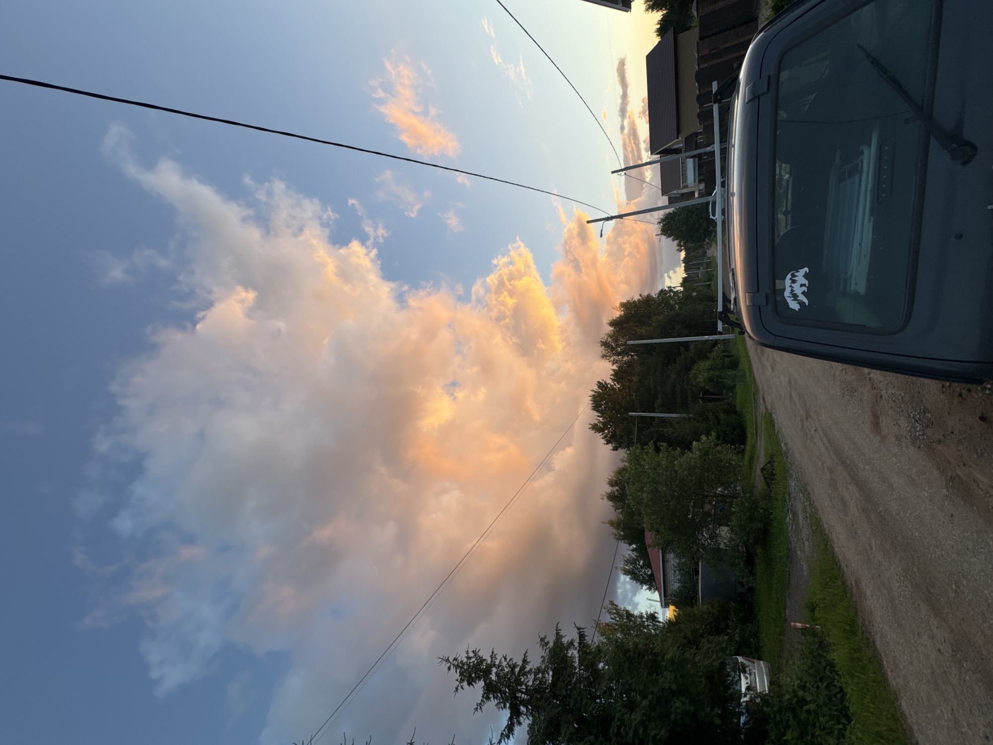A dark grey vehicle is parked on a dirt road with a line of trees and houses to the left. The sky is filled with dramatic clouds, lit with warm orange and pink hues from the setting sun. A black wire runs diagonally across the sky, connecting to utility poles that are partially visible. A white sticker of two bears is on the rear window of the vehicle. The scene evokes a peaceful, rural evening in Perm, Russia.
