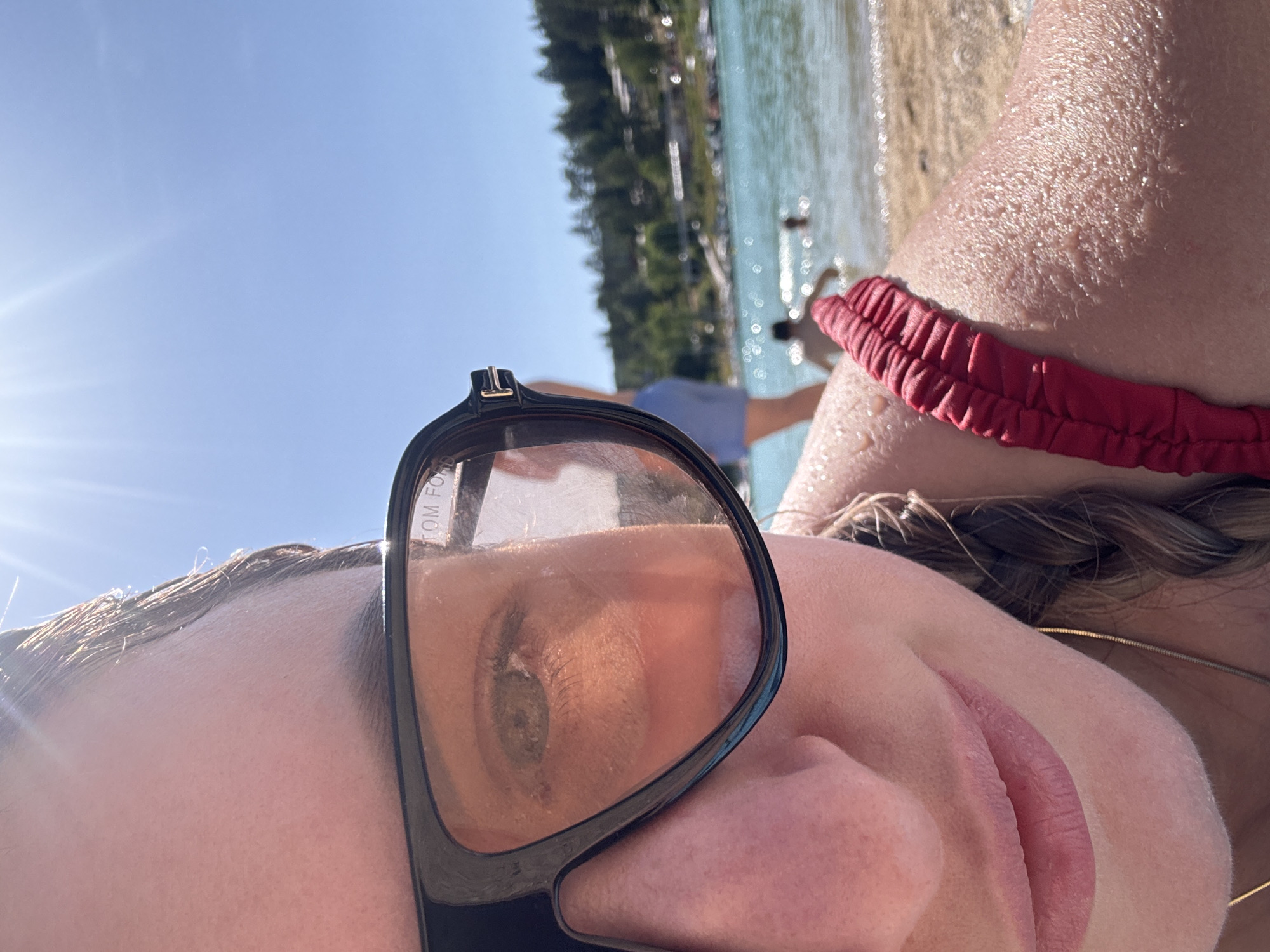 In Maisod, France, on a sunny day, a close-up shot focuses on the left side of a person's face, partially obscured by large black sunglasses with orange-tinted lenses. The person is wearing a red swimsuit strap across their shoulder. The background depicts a body of water, likely a lake, with people swimming and a shoreline lined with trees and buildings. The sky is a clear, bright blue with the sun glinting intensely, creating lens flare.