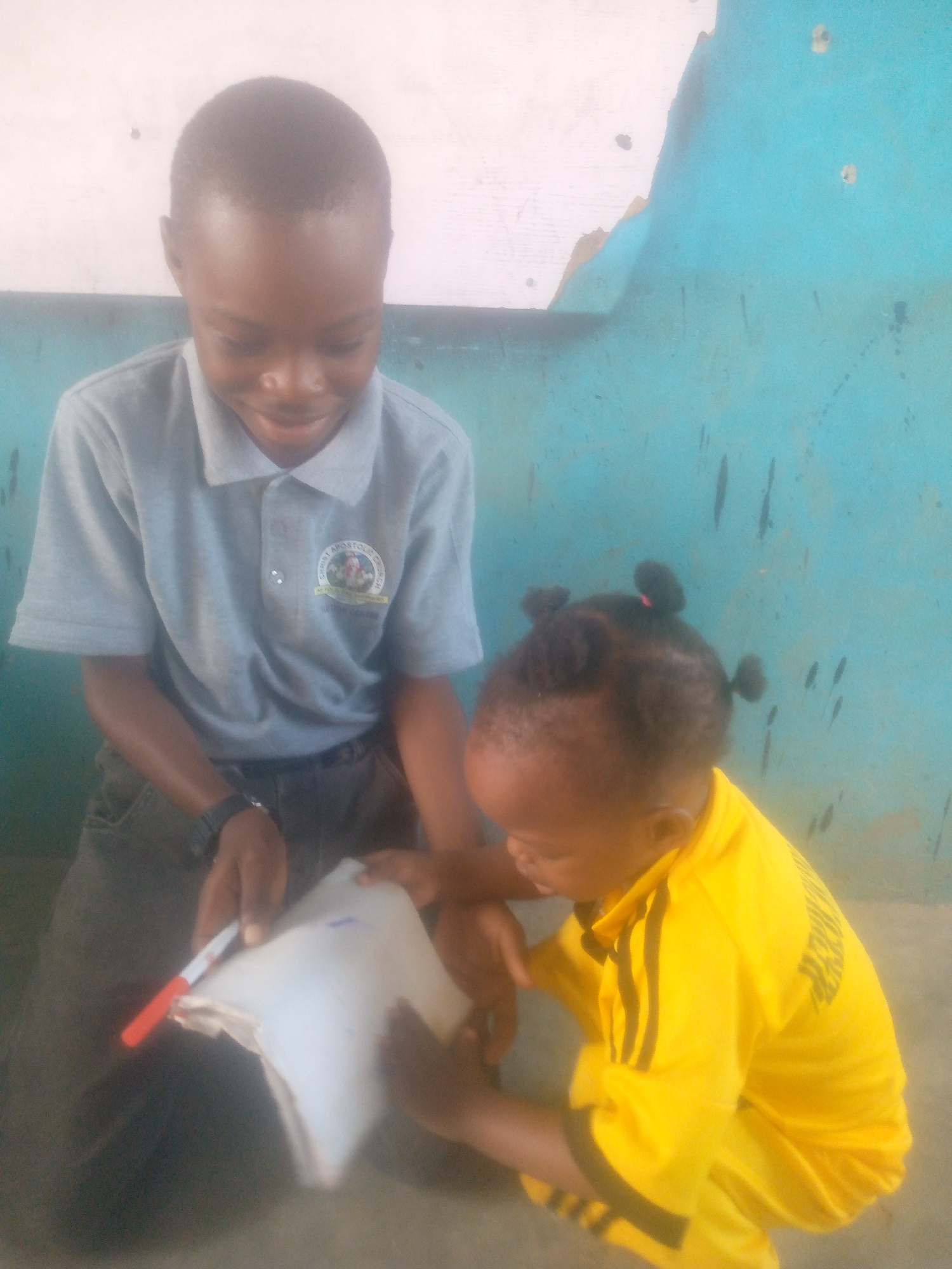 In an indoor setting in Unknown, Unknown, an older boy sits next to a toddler on the floor. The boy is dark-skinned with short hair and wears a grey collared shirt with a patch on the left side, dark pants, and a black wristwatch. He is holding a stack of white papers and a red marker. The toddler, also dark-skinned, is wearing a bright yellow long-sleeved shirt with black stripes on the sleeves and black pants. The toddler is looking at the papers in the boy's hands. The background consists of a wall with peeling paint, painted in shades of white and blue.