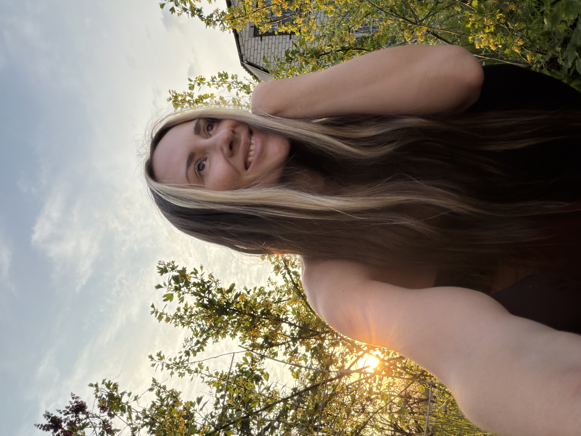 The image shows a woman with long brown and blonde hair smiling at the camera. She is outdoors, with trees and a building visible in the background. The sky is visible with a few clouds. The sun is shining, casting a warm glow on the scene. There is no precise location can be determined from this image.