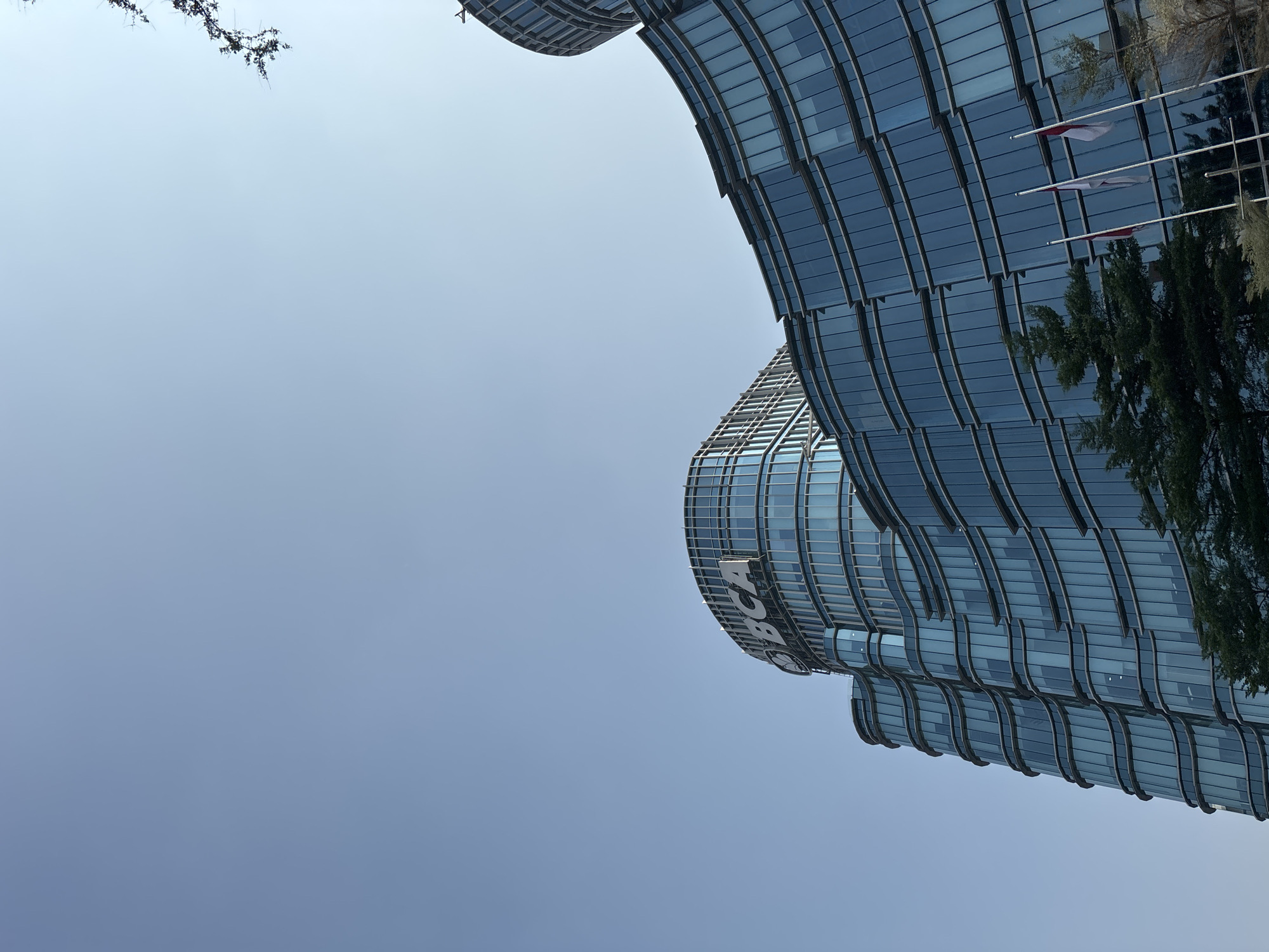 The image shows the BCA Tower in North Jakarta, Indonesia. It is a modern skyscraper with a curved glass facade. The building is tall and imposing, and it is set against a clear blue sky. There are trees and flags in front of the building, and the overall scene is one of urban prosperity and development. The BCA Tower is a prominent landmark in North Jakarta, and it is a symbol of Indonesia's economic growth. The building's unique design and its prominent location make it a popular tourist attraction.