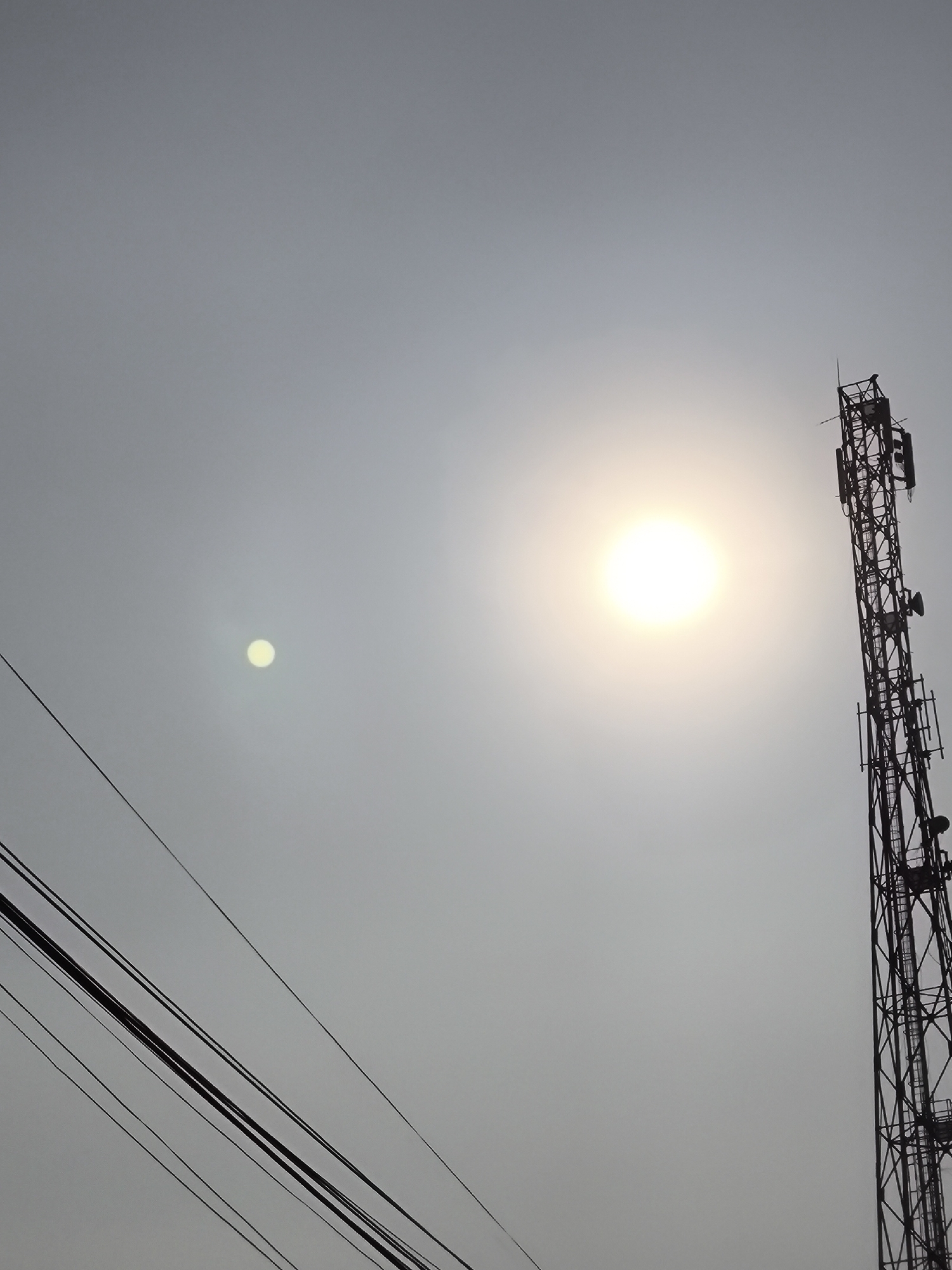 A cellphone tower is silhouetted against a hazy sky with the sun visible and glinting. The tower is located on the right side of the frame and is tall and metallic, with multiple antennas attached. In the foreground, on the left side of the image, are several power lines running diagonally across the frame. The sky is a uniform grey, suggesting a smoggy or overcast day. No people are visible in the image. The location is Cilegon, Indonesia.