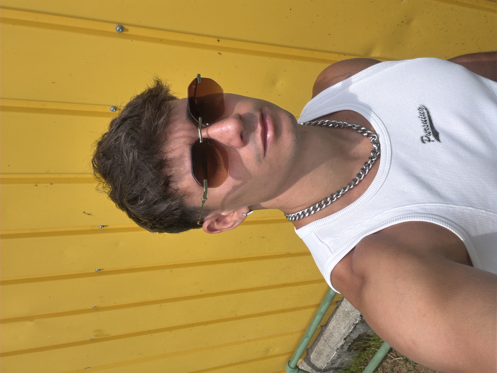 This media file features a close-up, vertically oriented shot of a young man posing against a bright yellow, corrugated metal wall. The man is wearing a white ribbed tank top and a thick, silver curb chain necklace. He has short, dark, messy hair and is wearing brown-tinted hexagonal sunglasses. He has an earring in his left ear. The image appears to be taken outdoors during the daytime, with natural light casting shadows and highlighting the textures of the wall and the man's skin. The bright yellow background suggests a sunny environment. A green pipe and some ground with grass are visible in the lower right corner, providing a hint of the outdoor setting. There is a small, embroidered logo on the left side of the tank top, which appears to read "Paradisio." The man's expression is neutral, and the overall mood is casual and confident. The location context is Sofia, Bulgaria.