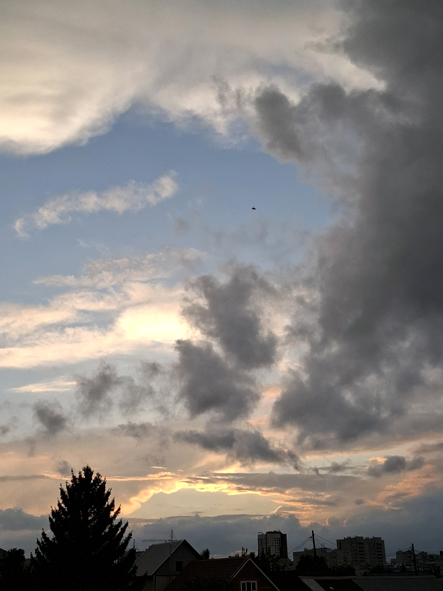 The image depicts a cloudy sky over a city, likely at dusk or dawn given the lighting. The sky is filled with various cloud formations, ranging from light, wispy clouds tinged with pink and yellow from the setting or rising sun, to darker, more ominous grey clouds on the right side of the frame. A silhouette of a tall evergreen tree stands prominently in the foreground on the left, partially obscuring the view of the city below. The cityscape consists of several buildings, including houses with visible rooftops and taller, multi-story structures in the distance. The presence of a lone, small dark object, possibly a bird or drone, is visible in the upper middle portion of the sky. The overall impression is one of a moody, atmospheric sky above an urban landscape. The context of Tyumen, Russia, suggests this scene is a typical view of the sky over that city, perhaps during transitional weather.