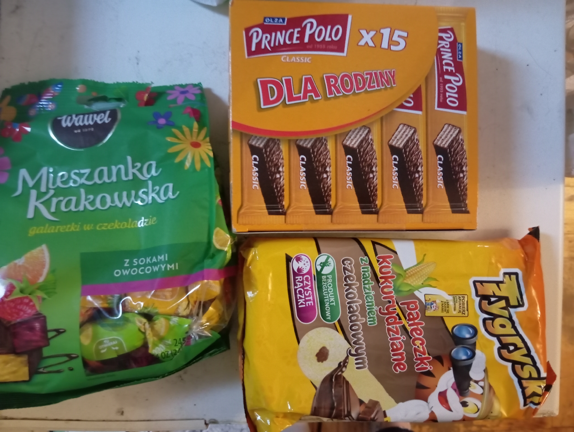 In the city of Zaporizhzhia, Ukraine, on a white, speckled surface that appears to be a shelf or countertop, three packages of snacks are arranged.
On the left, a large, bright yellow bag of "Tygrysky" chocolate-filled wafer rolls is positioned diagonally, with the top left corner of the bag closer to the viewer. The bag features an illustration of a cartoon tiger wearing binoculars and text in Polish.
In the center, a smaller, orange box contains individually wrapped "Prince Polo" classic wafer bars. The box is oriented vertically, and five wafer bars are visible, each displaying the "CLASSIC" label. The text "DLA RODZINY" and "X15" is prominently displayed on the orange packaging.
On the right, a green bag of "Mieszanka Krakowska" fruit-flavored jellies in chocolate is placed vertically. The bag is decorated with colorful flowers and contains visible individually wrapped jellies, some appearing to be lemon, raspberry, and orange flavored. Text in Polish indicates "Z SOKAMI OWOCWyMI" (with fruit juices).
The overall scene is a still life of confectionery items.