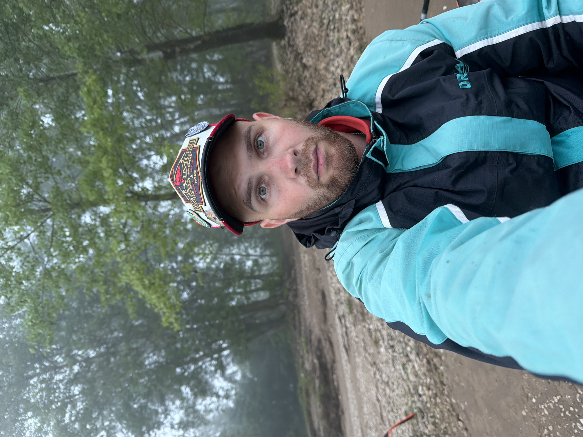 A man is depicted in a rotated close-up selfie, captured outdoors in Chaikovsky, Russia. He has a light beard and mustache, with light-colored eyes looking directly at the camera. He is wearing a distinctive cap, primarily white with red and black accents, featuring a prominent emblem on the front. Below the cap, he wears a black and teal/light blue outdoor jacket with white reflective stripes. A red collar is visible beneath his jacket.
The background shows a natural setting, likely a forest or wooded area, with numerous tall trees and green foliage, partially obscured by a significant amount of fog or mist, which gives the scene a soft, diffused light. A dirt or gravel path runs alongside him on the ground, which also appears stony. The man's expression is neutral or slightly serious.
Visible text on his cap reads "MAXIMUS THE BEST SPINNING RODS". On his jacket, the letters "DRA" are visible in teal stitching, likely part of a brand name such as "DRAGON". The overall impression is one of an individual in an outdoor, possibly wilderness, environment during a misty, overcast day.