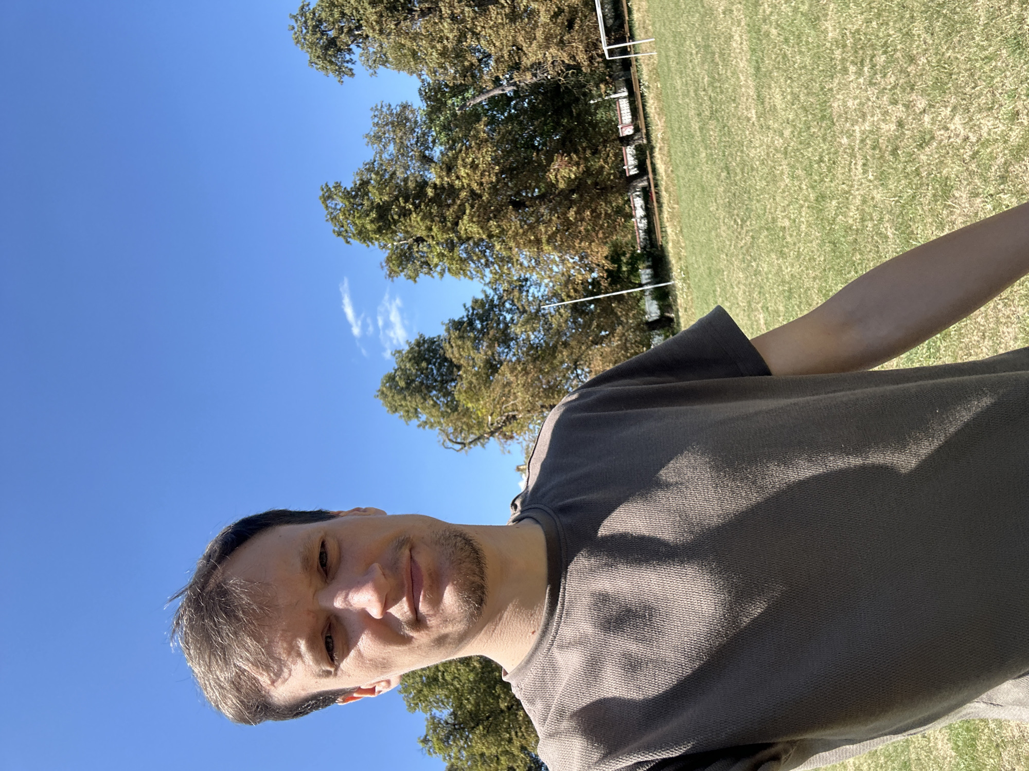 A man with short brown hair and a goatee is taking a selfie outdoors on a bright, sunny day. He is wearing a dark grey t-shirt. The background is a grassy field with several large trees and a clear blue sky with a few wispy clouds. A white goalpost is visible on the right side of the frame, suggesting this is a sports field. The location is specified as Makiv, Ukraine.