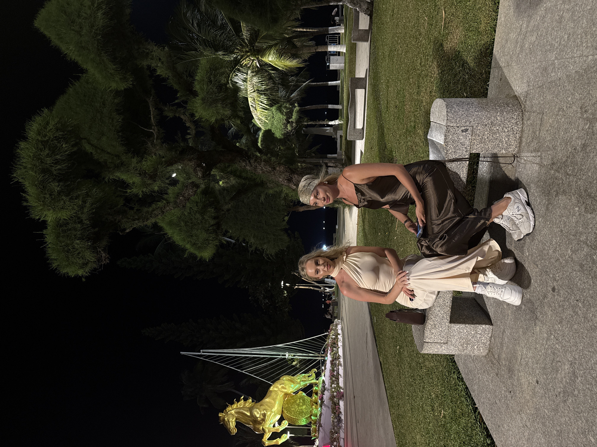 Two fair-skinned women are sitting on a stone bench at night in Nha Trang Ward, Vietnam. The woman on the left wears a light beige dress and white sneakers. The woman on the right wears a brown dress and white sneakers. Behind them is a park with trees and a golden horse statue with a bridge-like structure behind it. The area is lit by streetlights.