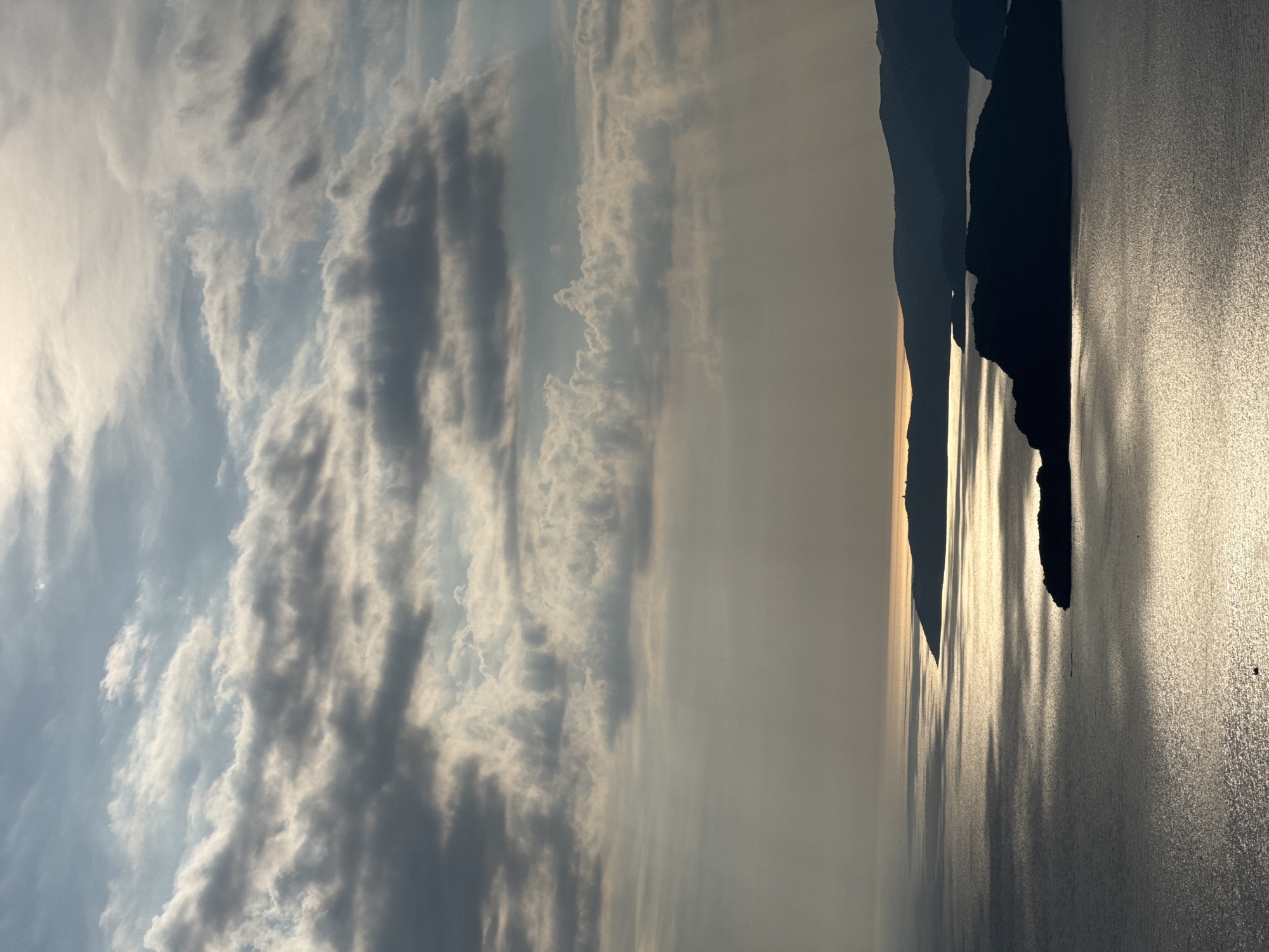 This is a photograph of the coastline near Sveti Stefan, Montenegro, taken from an elevated vantage point. The scene depicts the Adriatic Sea with several silhouetted islands and headlands. The sun is low in the sky, casting long, shimmering reflections on the water. Beams of light pierce through scattered clouds, creating a dramatic and atmospheric effect. The sky is a mix of blue and gray clouds, with patches of sunlight breaking through. The dark outlines of the landmasses contrast sharply with the bright reflections on the water. A small boat is visible on the sea in the lower left portion of the image. The overall impression is one of natural beauty and tranquility.