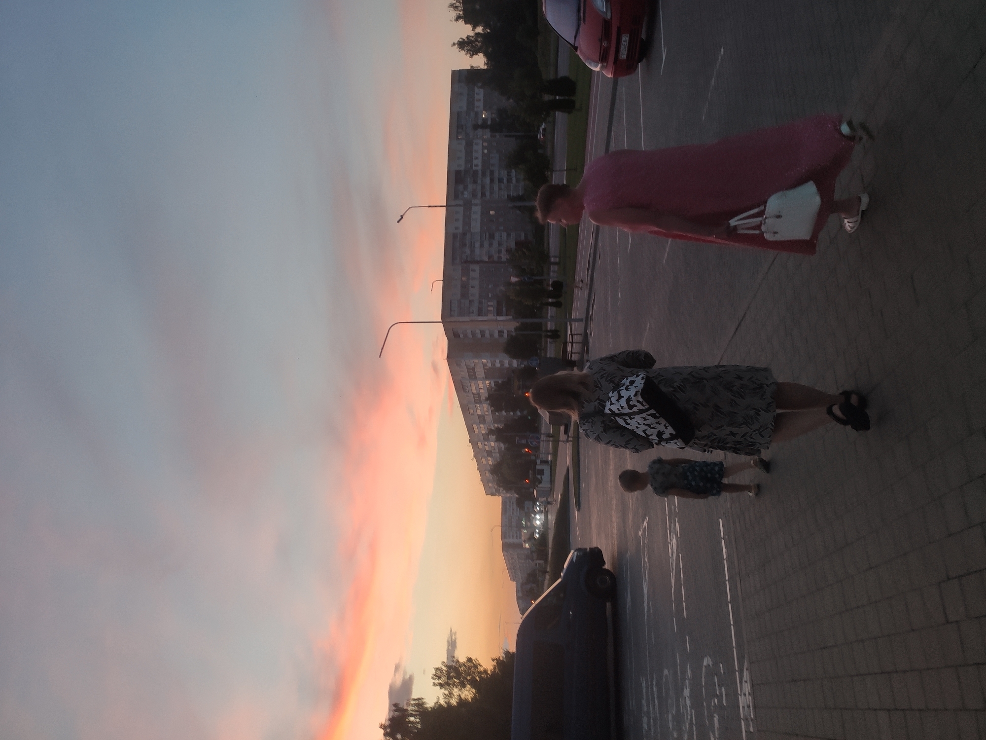 The image captures a street scene in Navapolatsk, Belarus, at dusk. A clear sky with streaks of pink and orange clouds stretches above a residential area with multi-story buildings. In the foreground, three figures are walking away from the camera on a paved walkway adjacent to a parking lot. On the left, a person in a dark-colored dress with a patterned backpack walks alongside a small child wearing a dark, patterned dress. To the right, another person in a long, flowing pink dress carries a white handbag. A dark-colored van is parked in the background of the parking lot, and a red car is visible on the right edge of the frame. Streetlights are illuminated in the distance, and trees line the area.