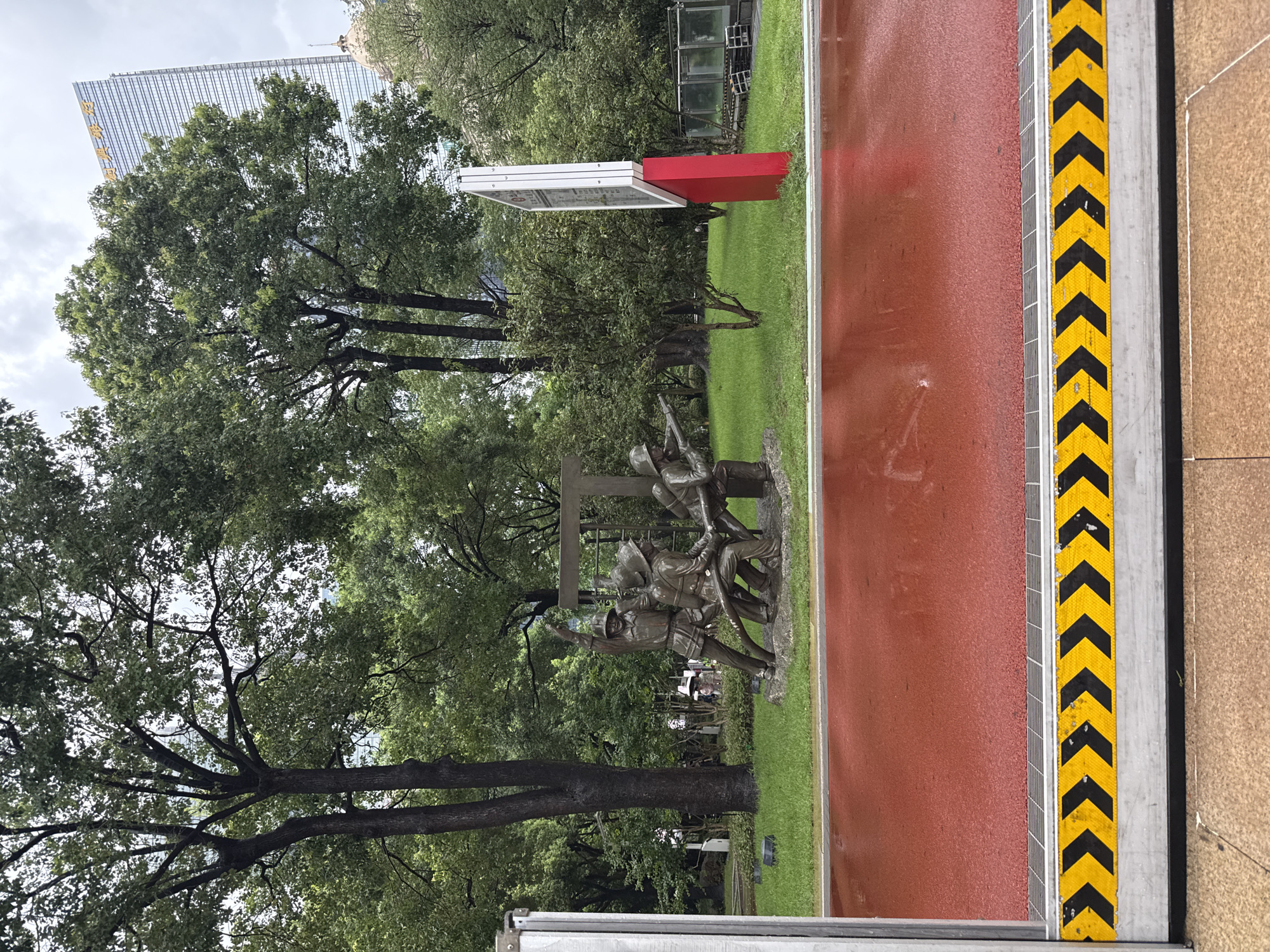 This image depicts a bronze sculpture of firefighters in action, located in Tianhe District, China. The sculpture, situated in a park-like setting, shows several figures working together, possibly holding a hose. The ground in front of the sculpture is a wet, red-colored surface, possibly a track or a paved area reflecting the overcast sky and surrounding trees. A building with a glass facade and golden lettering is visible in the background, towering above the trees. The foreground features a safety barrier with yellow and black chevron markings.