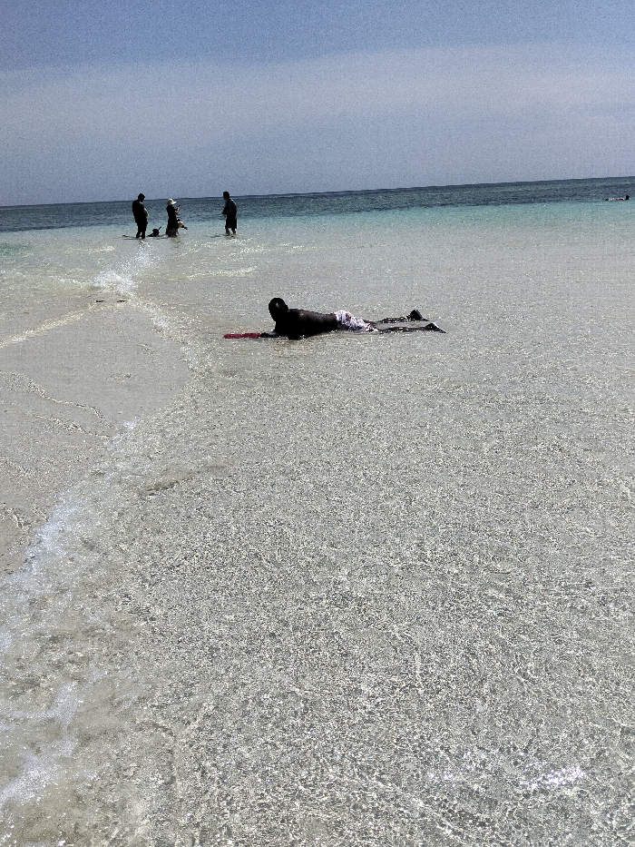 The scene is a shallow coastal area in Wasini, Kenya, characterized by clear, light turquoise water revealing a sandy bottom, transitioning to a deeper blue ocean under a pale blue sky. In the foreground, one dark-skinned individual is positioned prone in the shallow water, facing away from the viewer, wearing white and grey patterned swim shorts. A red boogie board or similar flotation device is situated directly in front of this individual. Approximately 20 meters further out, three additional individuals stand upright in the water. Two are dark-clothed figures; the third, centrally positioned, wears a light-colored hat. Their backs are largely oriented towards the camera. Minor wave activity is observable in the immediate foreground, creating a white foamy line. The horizon line is visible in the upper portion of the frame.