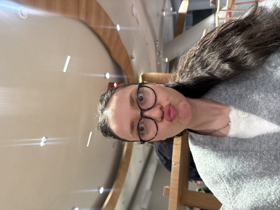 A woman with dark hair and glasses pouts at the camera. She wears a grey sweater over a white shirt. The background shows an interior space with a curved wooden beam and recessed lighting in the ceiling. She appears to be seated, with the back of a wooden chair visible. The photo is slightly tilted.