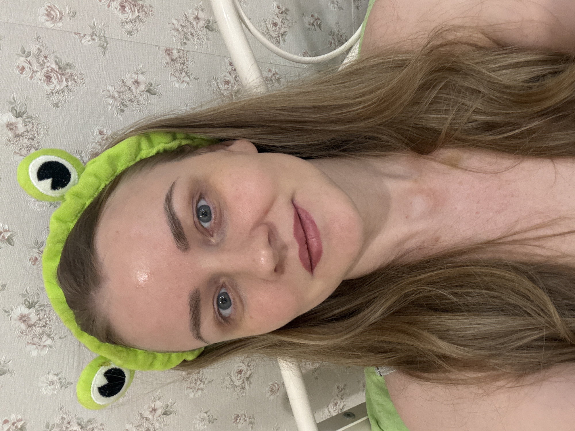 This is an angled, close-up portrait of a young woman, likely a selfie, taken indoors. The subject has light brown, wavy hair, blue eyes, and fair skin, looking directly at the camera with a soft, subtle smile. She is wearing a distinctive bright lime green frog-themed headband, featuring cartoon frog eyes on either side, which keeps her hair pulled back from her face. Her lips are lightly colored with a muted pink or mauve shade. A light green garment or strap is visible on her right shoulder, matching the headband's color palette.
In the background, a light-colored wallpaper with a repetitive floral pattern of muted pink roses and green leaves is visible, suggesting a bedroom or domestic setting. A white metal structure, possibly part of a bed frame or headboard, can be seen behind her. The lighting appears soft and natural, indicating daytime. The woman's expression conveys a calm and relaxed mood. No text is visible in the image. The setting is consistent with a personal space, possibly in Chaikovsky, Russia.