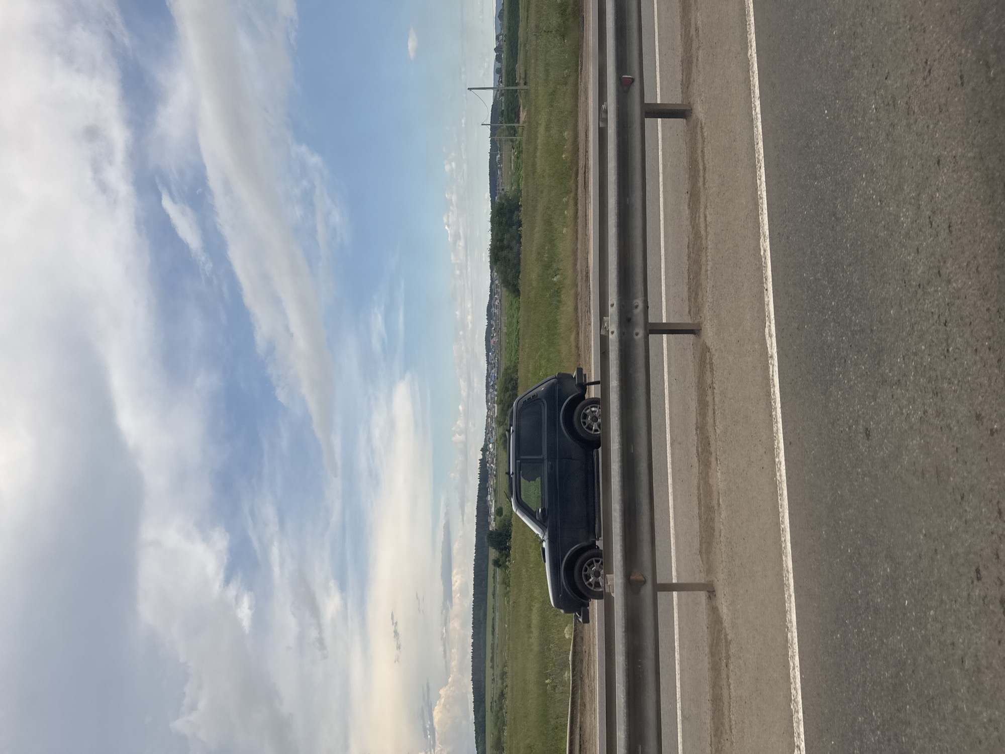 The image shows a dark-colored SUV, likely a Lada Niva, parked on the shoulder of a paved road in Kitsjanovo, Russia. The vehicle is positioned facing right in the frame, with its left side visible. The road has white lane markings and a metal guardrail runs along the right side of the frame. Beyond the road is a grassy field that stretches towards a treeline and a small village in the distance. The sky above is blue with scattered white clouds. The lighting suggests it is daytime, likely late afternoon or early evening, given the angle of the sun and the shadows. The weather appears to be fair. There is no text visible in the image.
