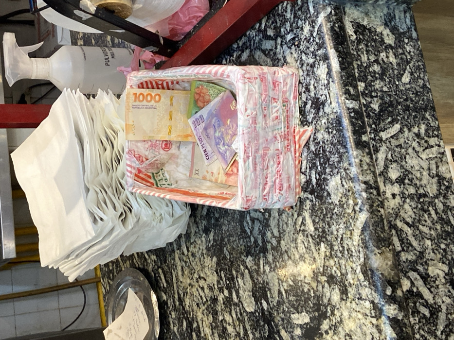 A small, striped box filled with Argentine pesos rests on a mottled gray countertop. Visible banknotes include a 1000 peso bill, a 100 peso bill, and other smaller denominations. The box sits adjacent to a stack of white paper bags and a white spray bottle. A small, silver dish with a receipt inside is also present. The scene appears to be a cashier area in a food establishment.