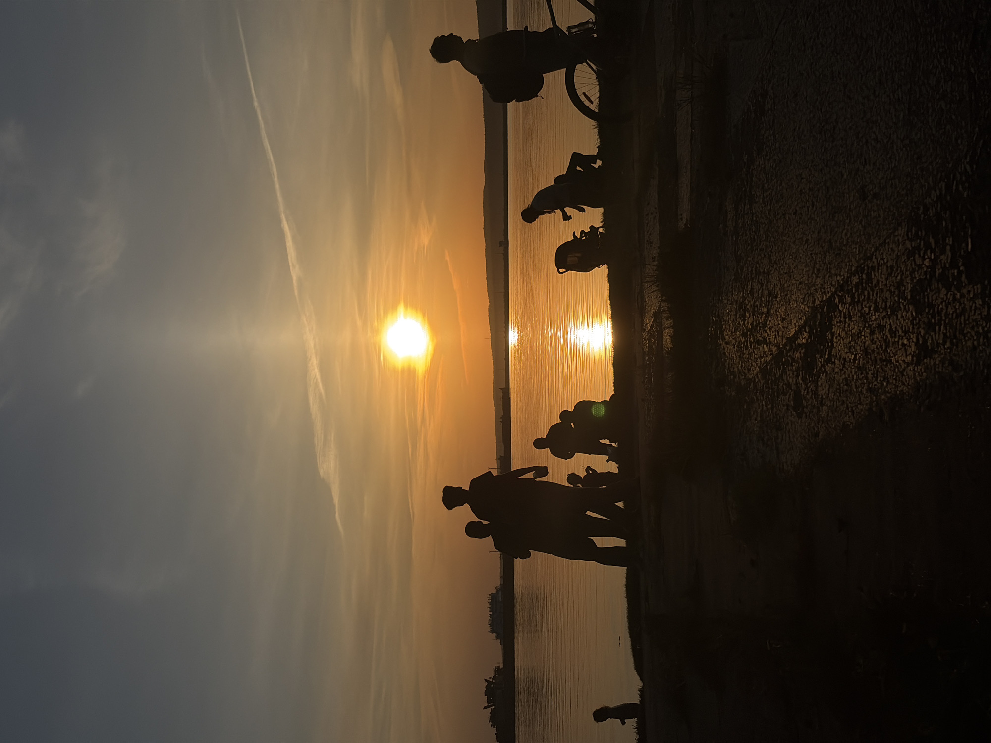The photograph depicts a sunset over the Kama River in Chaikovsky, Russia. Silhouetted figures of people are visible in the foreground, including adults and children. Some individuals are walking, and one person is on a bicycle. The sun is positioned low on the horizon, reflecting brightly on the water's surface. Contrails are visible in the sky above. The area appears to be a paved walkway or embankment alongside the river.