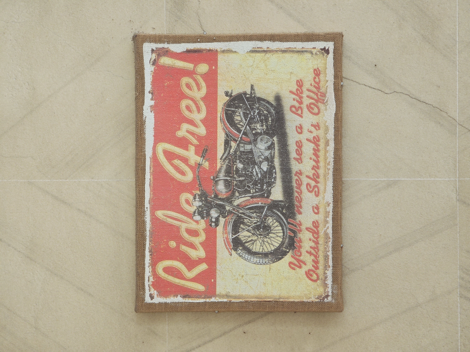 The image shows a rectangular canvas with a vintage-style depiction of a motorcycle and text. The text reads "Ride Free!" at the top and "You'll never see a Bike Outside a Shrink's Office" at the bottom. The canvas is framed by a rough, tan material, possibly burlap. The canvas is pinned to a light-colored, tiled wall. The tiles are large and have a subtle pattern. The image is well-lit, with the light source appearing to be overhead. There are no people visible in the image. The scene appears to be indoors, with the wall suggesting an interior space. The overall aesthetic is retro and perhaps humorous. The location is identified as Xiaoshan District, China, although there are no specific visual cues in the image to confirm this.