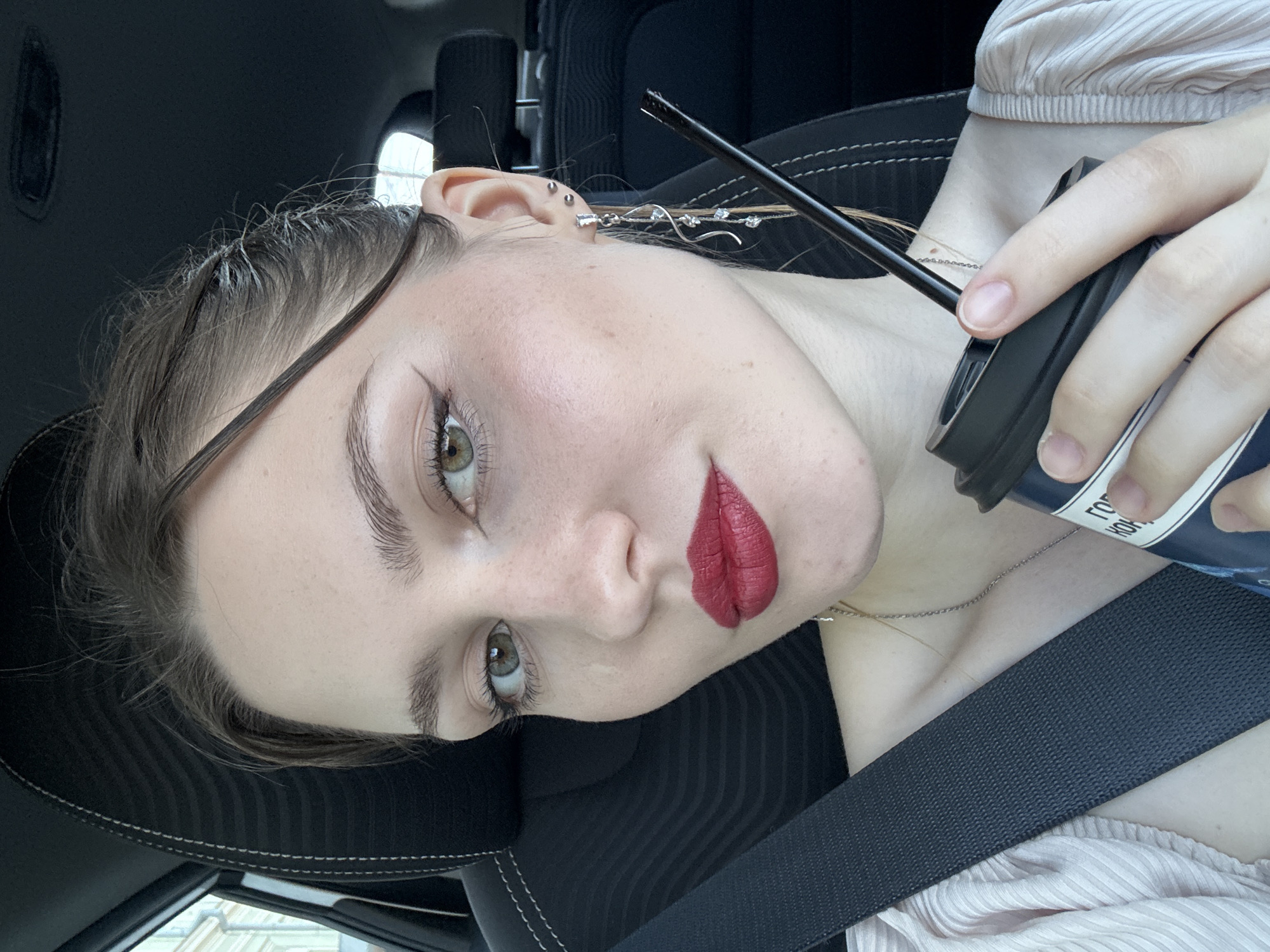 A young adult female is captured in a close-up selfie from inside a vehicle, positioned in the city of Saint Petersburg, Russia. She has light skin, green eyes with winged black eyeliner, and red lipstick. Freckles are visible across her nose and cheeks. Her dark brown hair is pulled back, with some wisps framing her face. She wears a light-colored, possibly pale pink, pleated top and a delicate silver-toned chain necklace. On her right ear, she has a long, dangling silver-toned earring adorned with multiple clear crystals, along with two small stud piercings higher on the earlobe. Her right hand, with manicured nails, holds a dark blue/black disposable coffee cup with a black lid and straw. Visible white Cyrillic text on the cup reads "ГОРЬКИЙ КОФЕ" (Bitter Coffee). She is secured by a dark seatbelt across her chest. The interior of the vehicle features a black car seat with white stitching on the headrest and a dark ceiling. Through the left window, a light-colored, multi-story classical stone building characteristic of European urban architecture is partially visible.