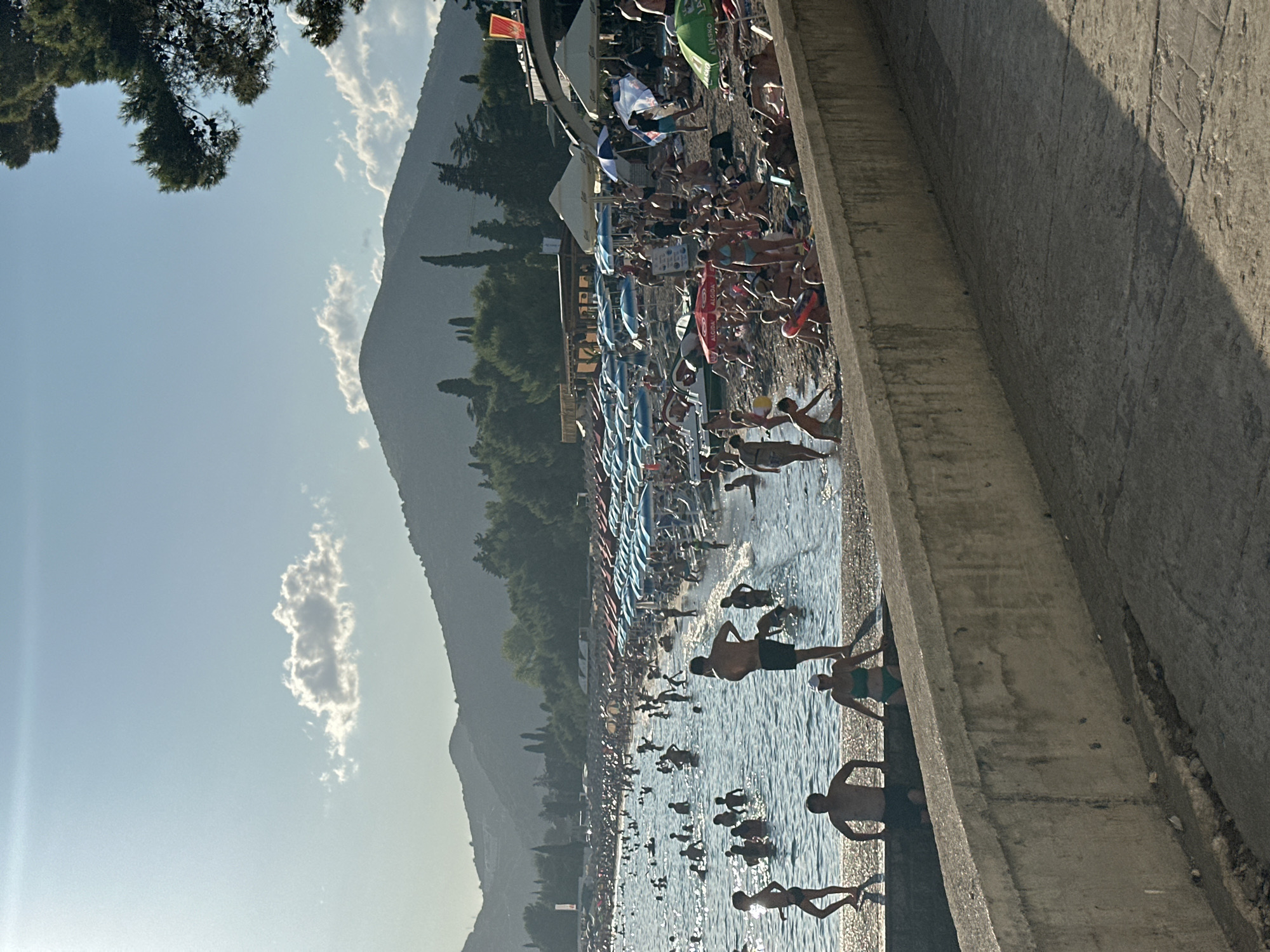 This image captures a crowded beach scene in Budva, Montenegro, under a clear sky with sparse clouds. The foreground features a concrete wall and walkway casting shadows, indicating it's late afternoon. Beyond the wall, a busy beach stretches along the coastline. Numerous people are visible, with many in the water, swimming or wading. Others are on the pebbled shore, lying on beach chairs, or standing under colorful umbrellas. The water is calm and appears to be the Adriatic Sea. In the background, lush green trees and rolling hills rise, framing the beach and hinting at the mountainous landscape of Montenegro. The overall atmosphere is one of a vibrant summer day at a popular tourist destination.