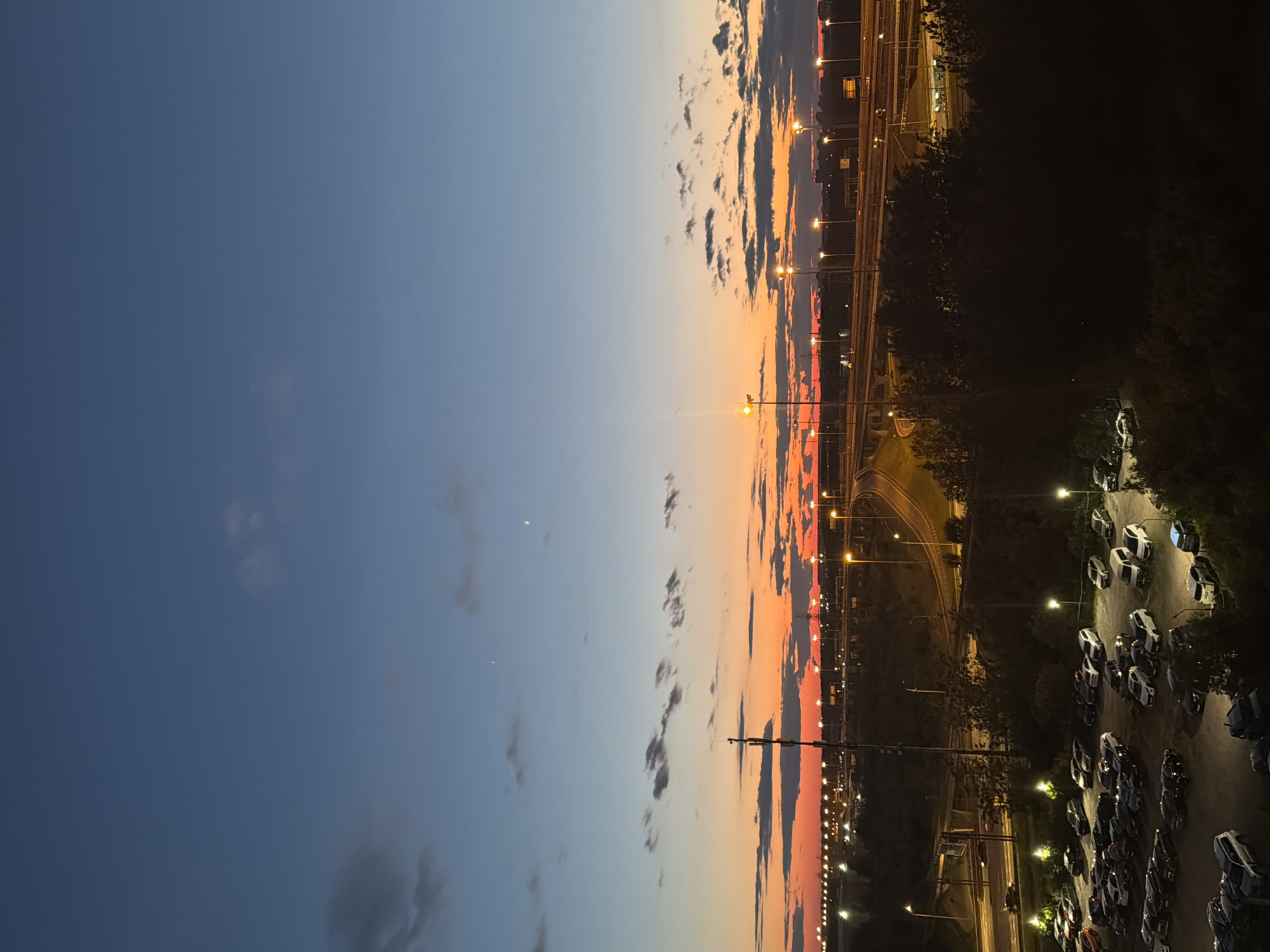 A high-angle, wide shot captures a twilight sky over a city. The sky transitions from a deep blue at the top to a vibrant orange and red near the horizon, with scattered clouds. A multi-lane highway, illuminated by streetlights, curves through the scene. In the foreground, a parking lot filled with cars is visible, with some vehicles being lit by overhead lamps. Trees and other foliage dot the landscape, creating a sense of depth. The overall impression is one of an urban environment at dusk.
