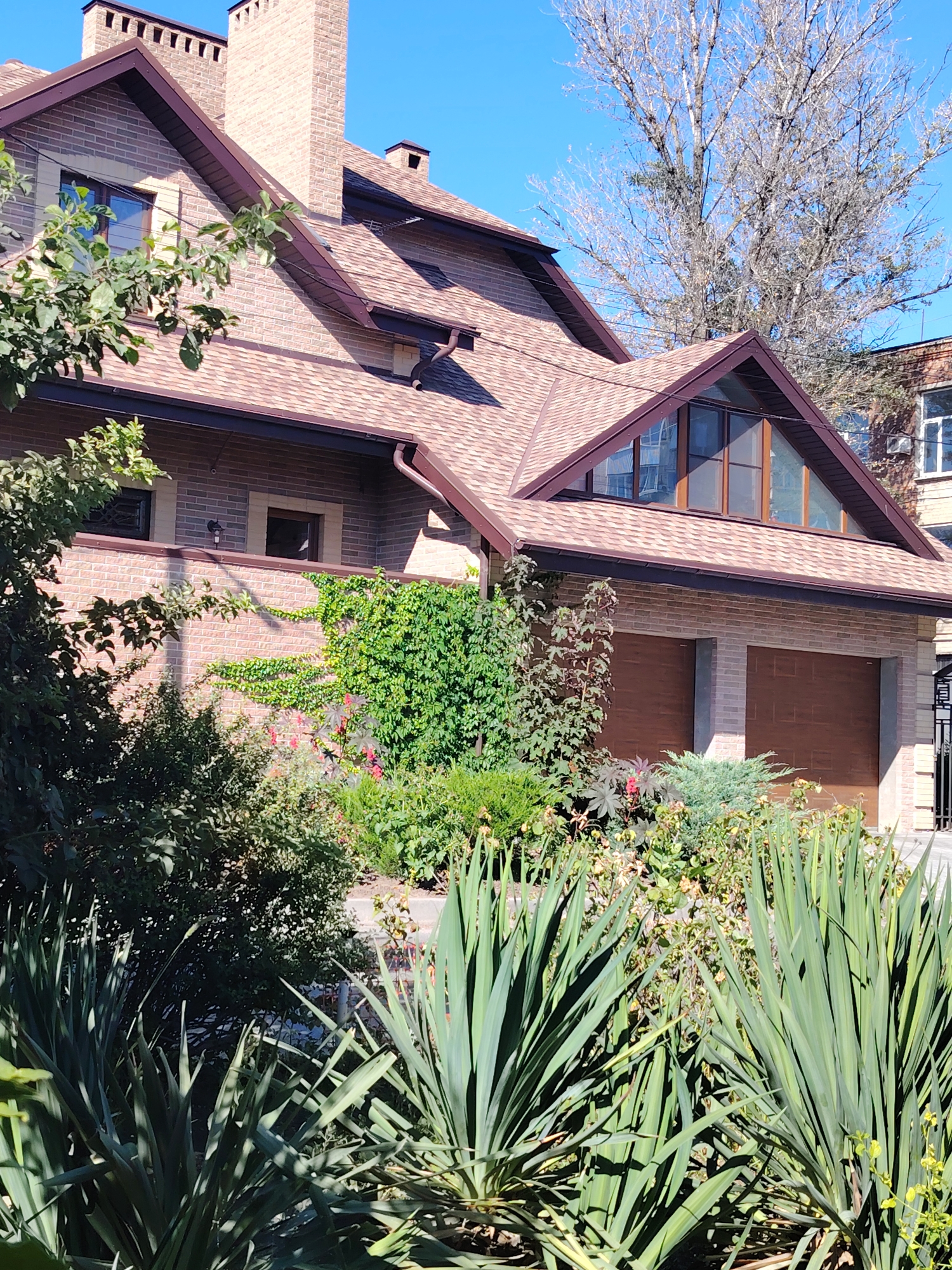 The image shows the facade of a multi-story brick house with a brown tiled roof. Two garage doors are visible on the ground floor. The house is surrounded by lush greenery, including trees, bushes, and flowering plants. The sky is clear and blue. The image was taken on a sunny day. This location is believed to be in Rostov-on-Don, Russia, based on the architectural style and vegetation, which are typical for the region.