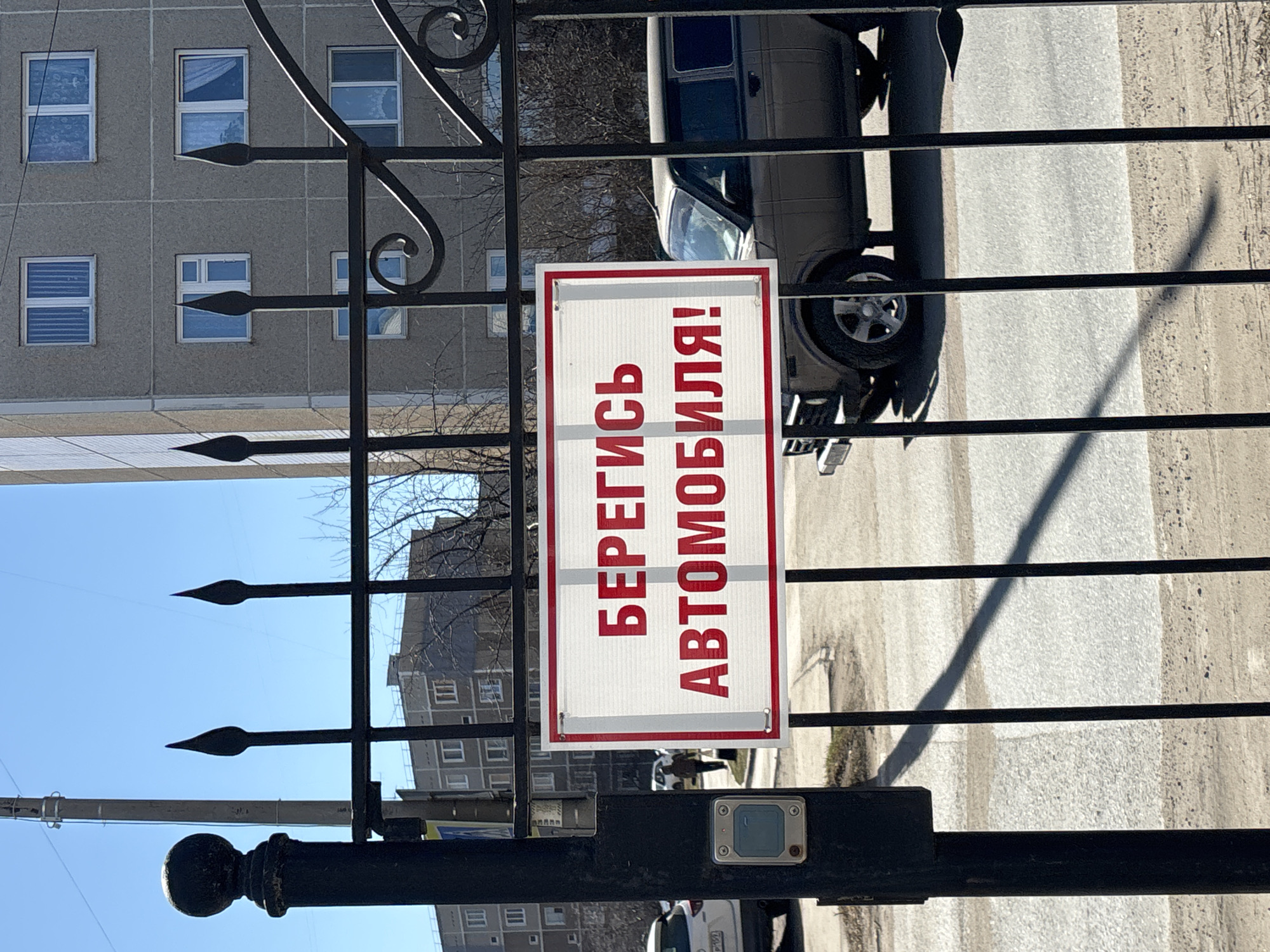 The image shows a black wrought iron gate with a sign attached to it. The sign is rectangular with a white background and a red border. The text on the sign is in Russian, written in red letters, and translates to "Beware of the car!". Behind the gate, there is a view of a city street in what is stated to be Chaikovsky, Russia. A light-colored van is parked on the street. In the background, there is a multi-story grey building with several windows. The sky is blue. The gate includes spear-shaped finials and decorative scrolls.