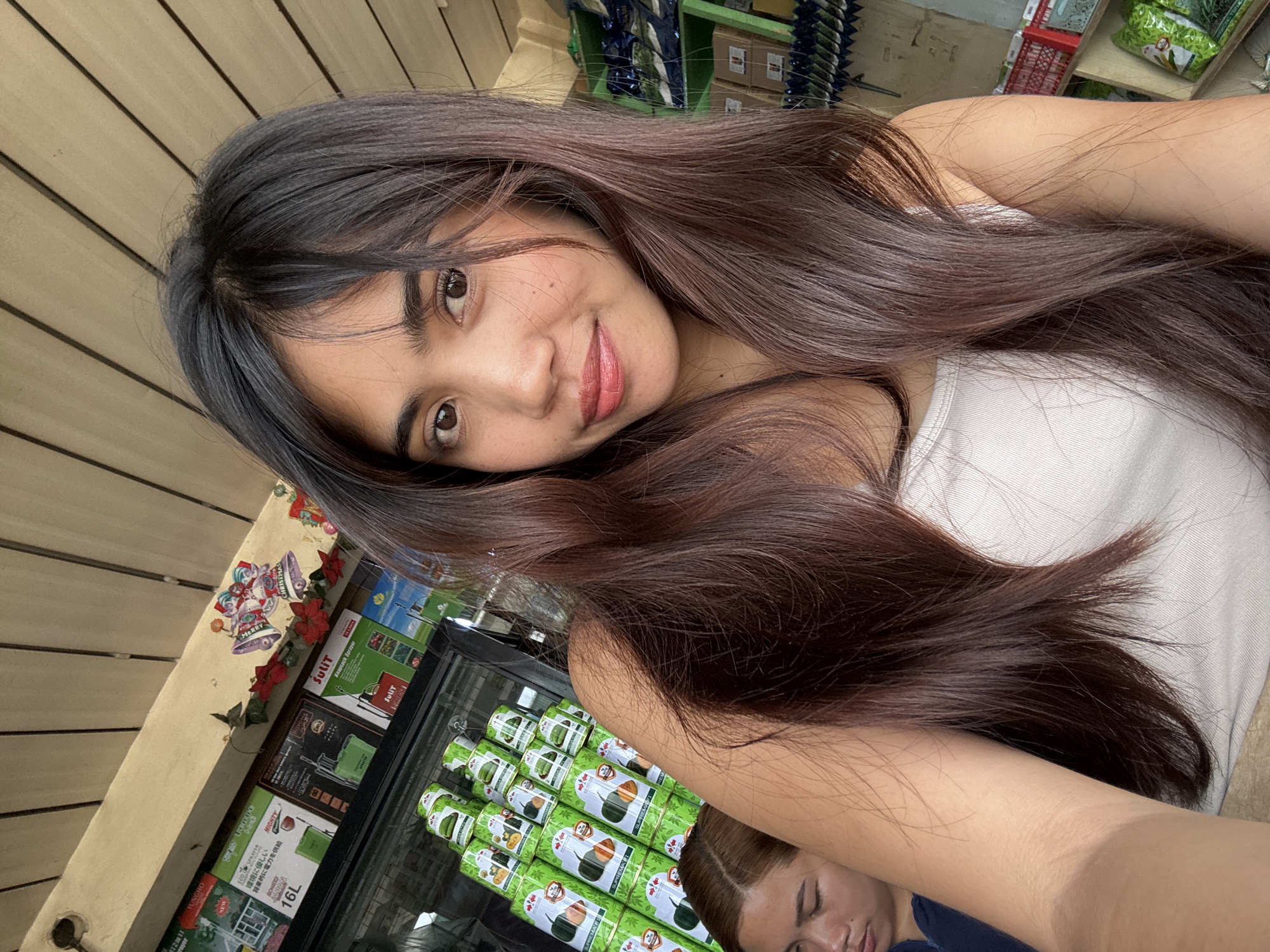The image is a selfie taken by a young woman with long, dark brown hair. She is wearing a white tank top and is positioned in the foreground. To her left and slightly behind her, another woman with brown hair is visible, looking downwards. The background suggests an indoor environment, possibly a store or market, with shelves stocked with various items. Prominently displayed are stacks of green cans with white labels featuring illustrations of food items, and above these, there are various boxes and posters on the wall. Christmas decorations, including a banner that says "Merry Christmas" and figurines of Santa Claus, are visible on the upper part of the wall. The ceiling appears to be made of wooden planks. The overall setting is casual and appears to be a commercial space.
