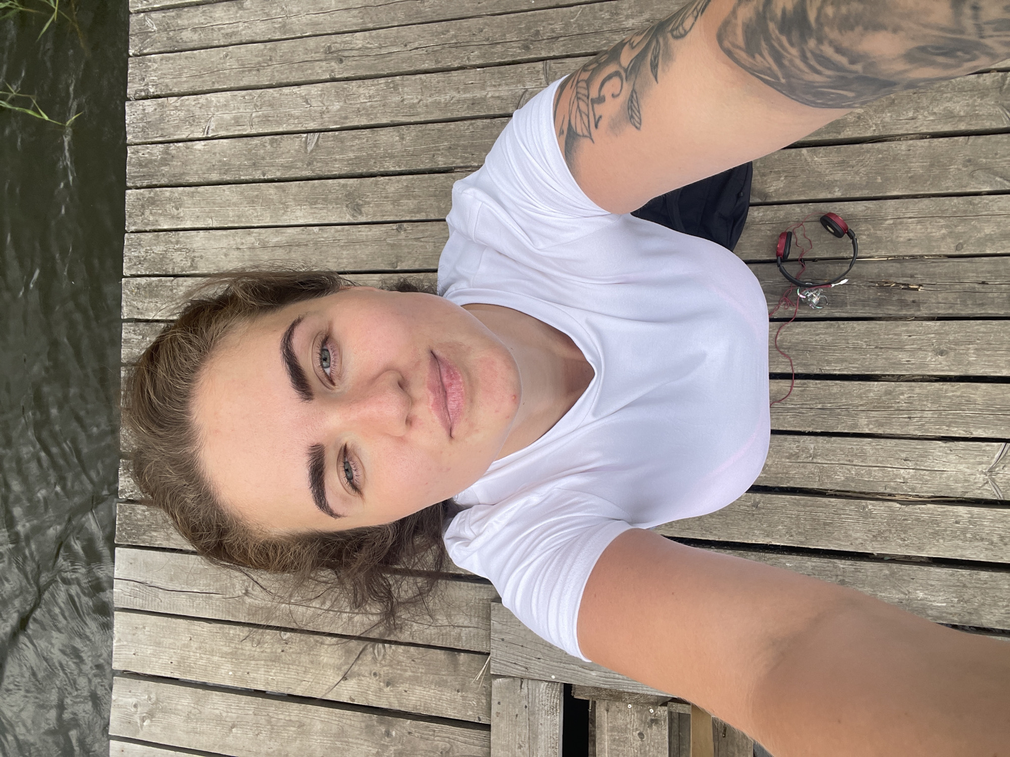 A young woman with brown hair and light blue eyes is lying on a wooden dock next to a body of water. She is wearing a white t-shirt and has tattoos on her arm. There is a pair of red headphones on the dock near her. The location is the city of Ostrov, Russia.