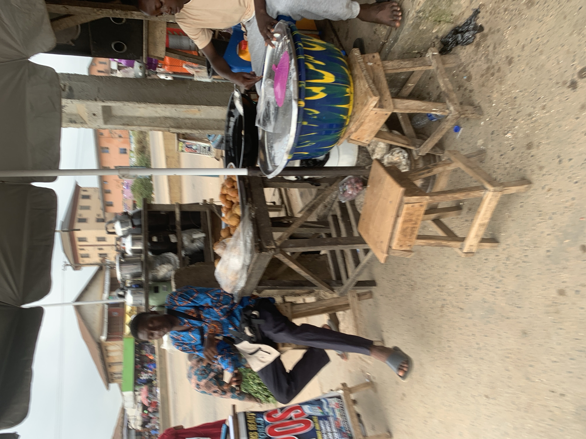 A street vendor is seen in Shagamu, Nigeria, preparing food outdoors under a large umbrella. The man, wearing a blue and orange patterned dashiki and dark pants, sits on a stool to the left, with a beige messenger bag slung across his chest. In front of him, a table displays various items, including what appear to be fried snacks and a stack of metal pots on a shelf behind. To the right, another person, wearing a light-colored t-shirt and grey sweatpants, is actively engaged in food preparation, working with a large blue and yellow patterned basin filled with water. The ground is a dusty, sandy surface, and the background shows buildings and other street activity, indicating a busy urban environment. There is a sign with the text "KS BEAUTY WORLD POS" visible on the left side of the frame.